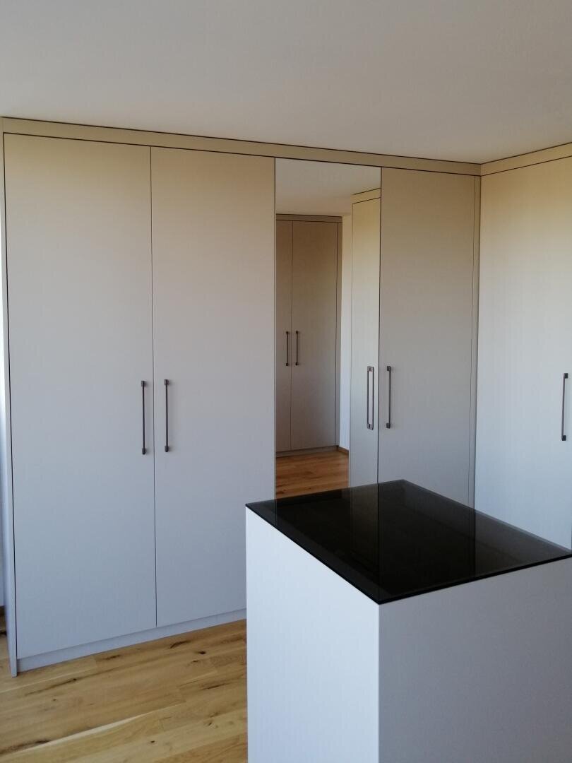 Minimalistisches Schlafzimmer mit großen weißen Einbauschränken und hohen schwarzen Griffen, hellem Holzfußboden und einer zentralen weißen Insel mit einer glänzenden schwarzen Oberfläche. Der Raum ist gut beleuchtet und übersichtlich.