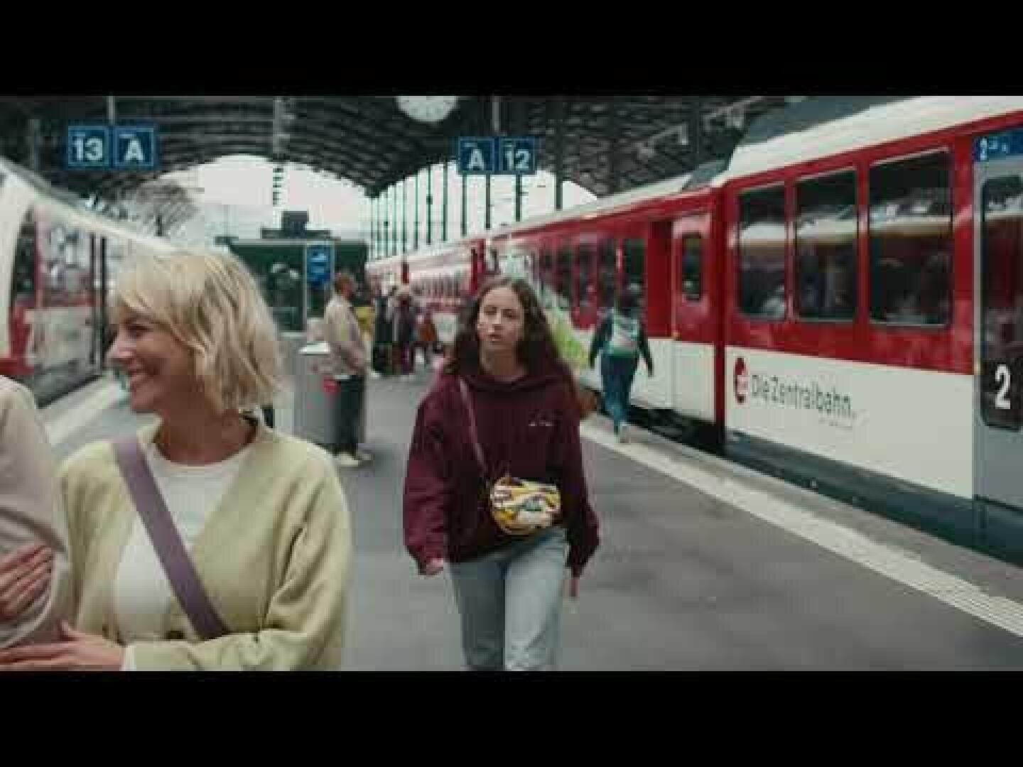 Eine Frau in einem kastanienbraunen Kapuzenpullover geht auf einem Bahnsteig zwischen zwei rot-weißen Zügen, während eine andere Frau in einem hellen Pullover lächelt und nach links schaut. Die Schilder darüber zeigen die Bahnsteignummern A12 und A13.