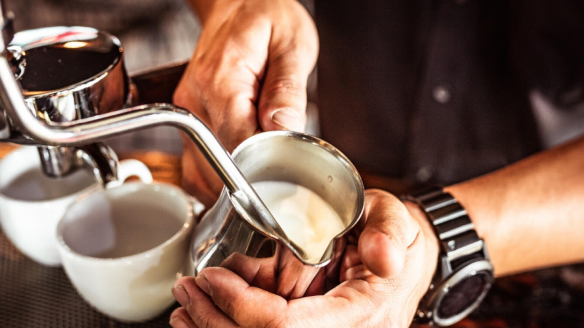 Eine Person, die eine Uhr trägt, brüht Milch in einer Metallkanne mit einer Espressomaschine auf, während zwei weiße Kaffeetassen in der Nähe auf dem Tresen stehen.