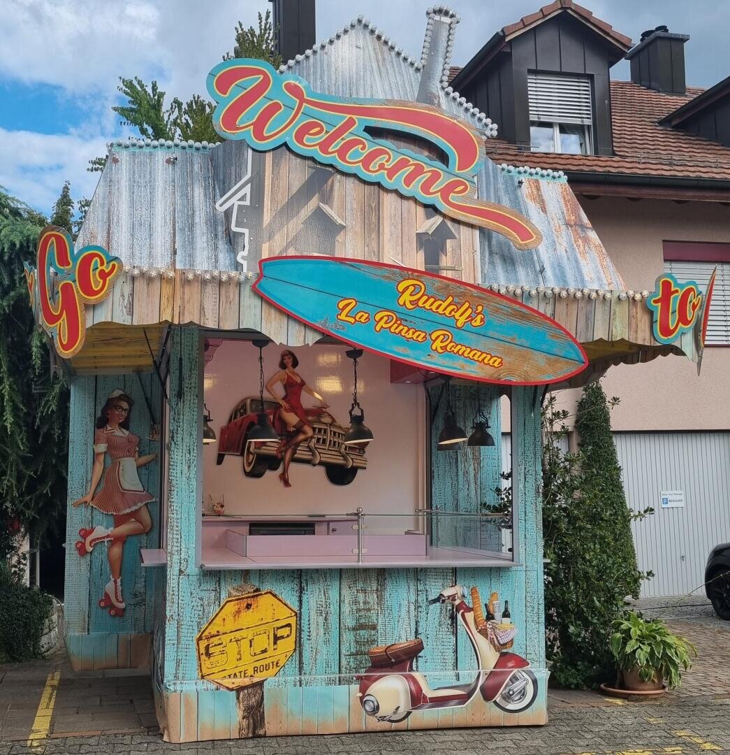 Ein farbenfroher Lebensmittelkiosk im Retro-Stil mit Vintage-Schildern, Bildern von Frauen in Kleidung der 1950er Jahre und einer Vespa-Illustration. Auf dem Schild steht Rudys La Pizza Romana. Der Kiosk ist von Häusern und Grünanlagen umgeben.