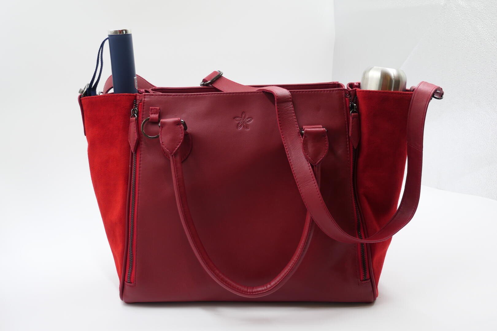 Eine große rote Tragetasche mit zwei Henkeln und einem Schulterriemen, mit Seitentaschen für eine blaue Flasche auf der linken und eine silberne Flasche auf der rechten Seite, vor einem schlichten weißen Hintergrund.