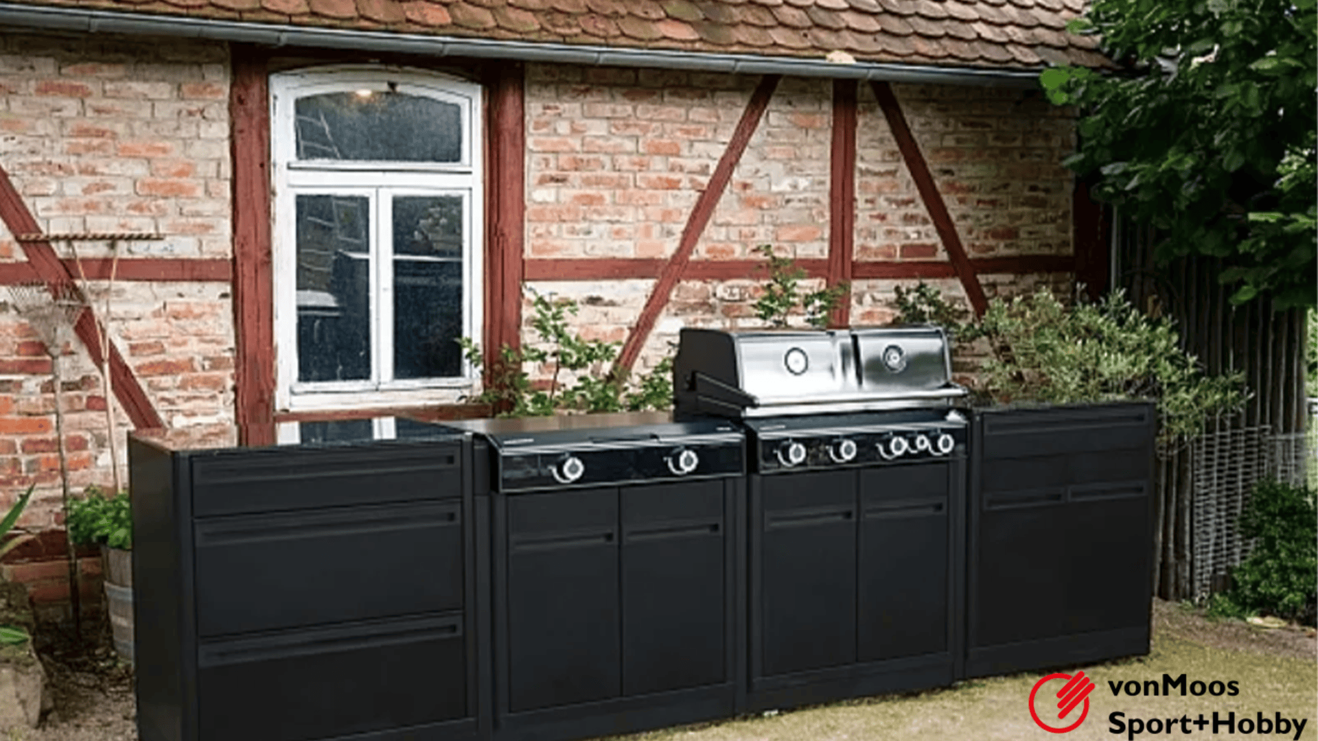 Outdoor-Kücheneinrichtung mit schwarzen Schränken und einem silbernen Grill vor einem rustikalen Backstein- und Holzhaus, mit Pflanzen und einem Fenster im Hintergrund. vonMoos Sport+Hobby-Logo in der unteren rechten Ecke.
