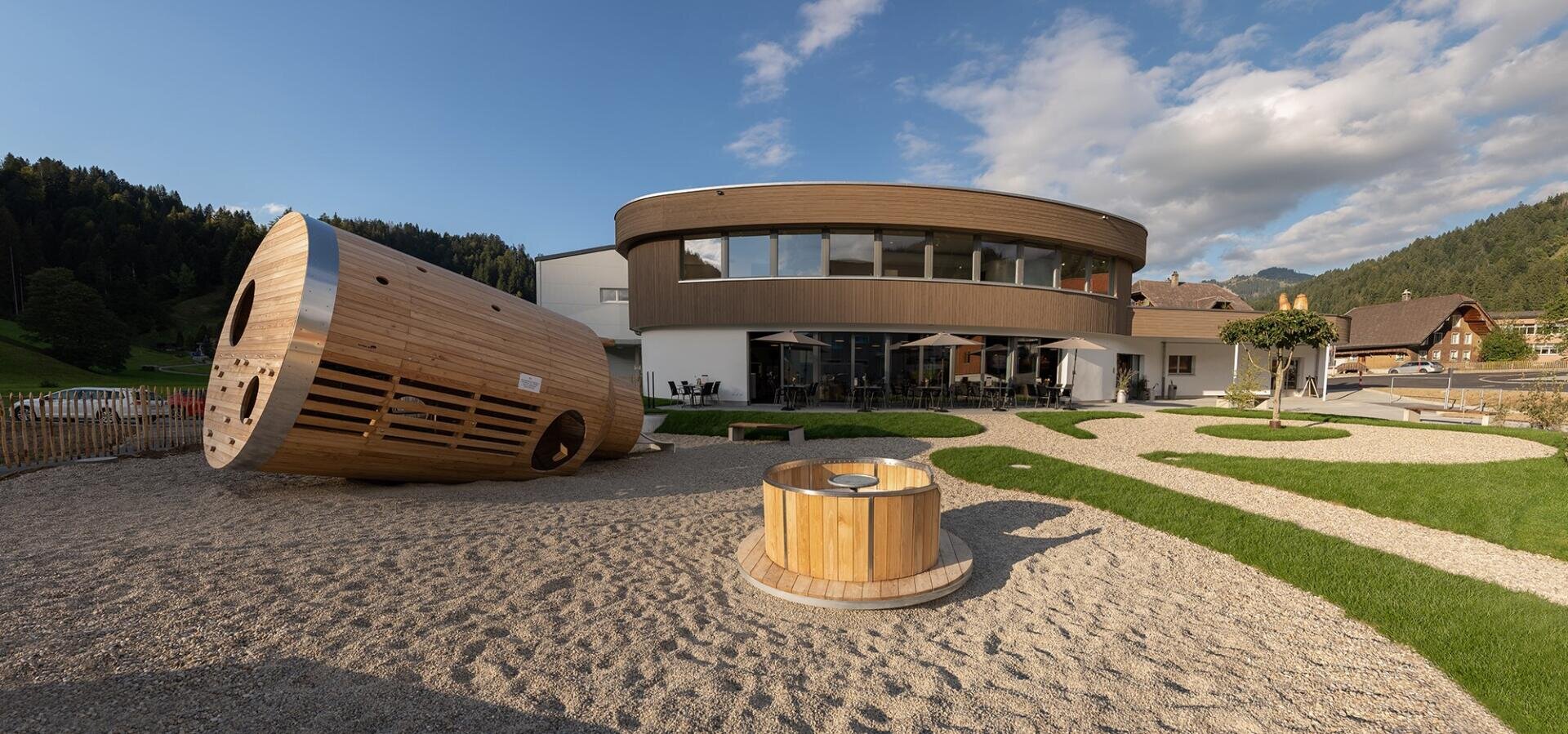 Ein modernes Gebäude mit rundem Obergeschoss und großen Fenstern befindet sich hinter einem Spielplatz mit einer großen hölzernen Tunnelkonstruktion und einer runden Bank auf Kies, umgeben von grünem Gras und Bergen.