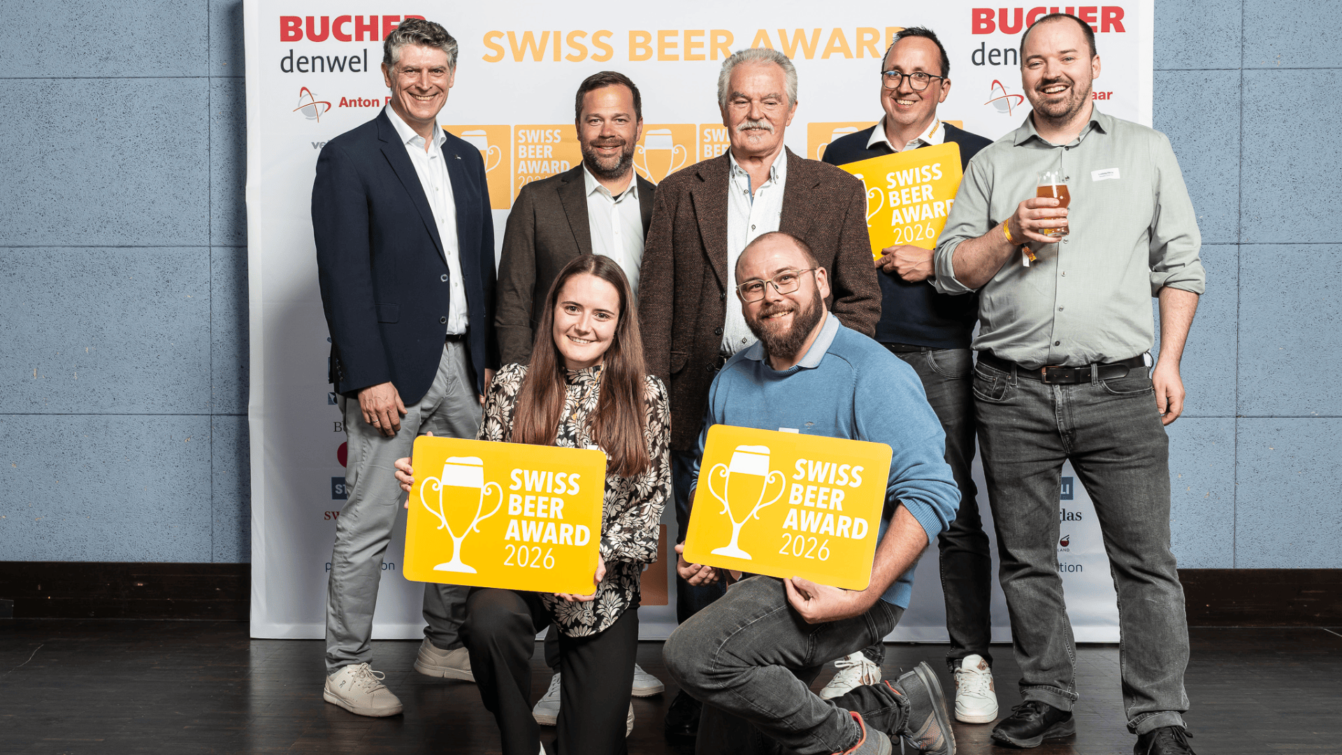 Sieben Personen posieren und lächeln an der Swiss Beer Award-Veranstaltung und halten gelbe Schilder mit der Aufschrift Swiss Beer Award 2026. Hinter ihnen befindet sich eine Kulisse mit Sponsorenlogos. Eine Person hält ein Glas Bier in der Hand.