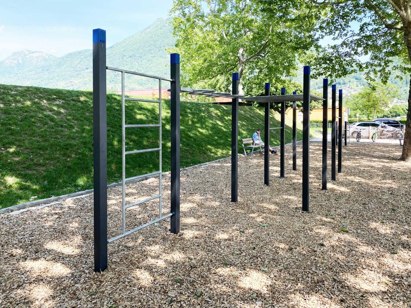Outdoor-Fitness-Park Struktur mit Monkey Bars und eine Leiter, auf Holzspänen unter belaubten Bäumen platziert. Eine Person sitzt auf einer Bank im Hintergrund, mit einem grasbewachsenen Hügel und Bergen sichtbar.