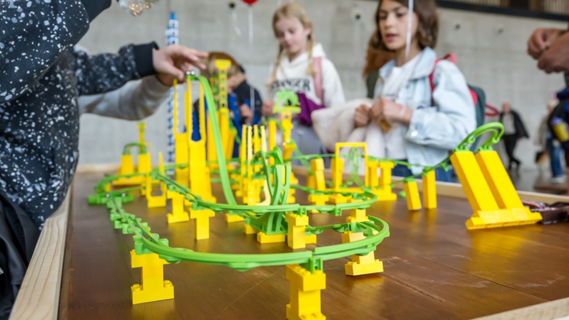 Kinder versammeln sich um einen Tisch und untersuchen und spielen mit einem bunten Konstruktionsspielzeug mit einer gewundenen grünen und gelben Achterbahnschiene. Der Hintergrund zeigt eine graue Wand und weitere Personen, die das Geschehen beobachten.