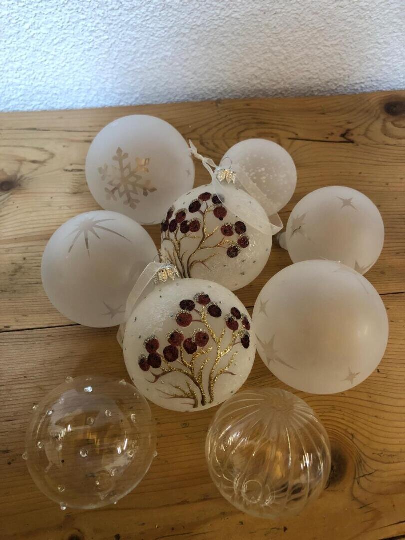Eine Gruppe von Weihnachtsschmuck aus Glas, größtenteils mattiert mit geätzten Motiven, umgibt zwei zentrale Ornamente, die mit roten Beeren und goldenen Zweigen verziert sind, alle auf einer hölzernen Oberfläche angeordnet.