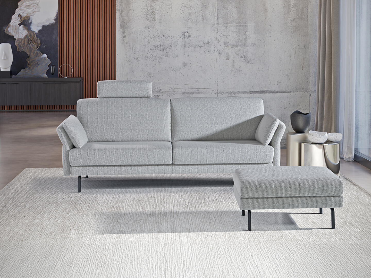 Ein modernes Wohnzimmer mit einem hellgrauen Sofa, einer passenden Ottomane, einem strukturierten Teppich und einer minimalistischen Einrichtung mit Vasen und abstrakter Kunst vor einer Betonwand und Holzverkleidung.