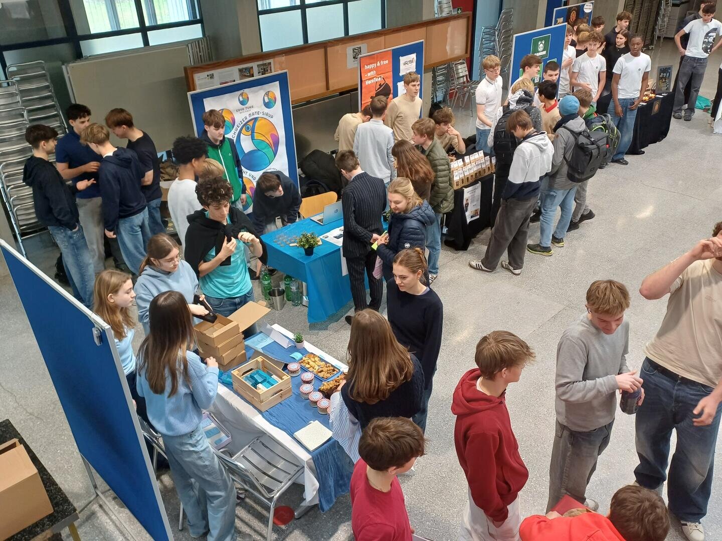 Eine Gruppe von Schülerinnen und Schülern versammelt sich bei einer Indoor-Veranstaltung oder Schulmesse um mehrere Ausstellungstische mit Postern, Snacks und Produkten, unterhält sich und erkundet die Stände.