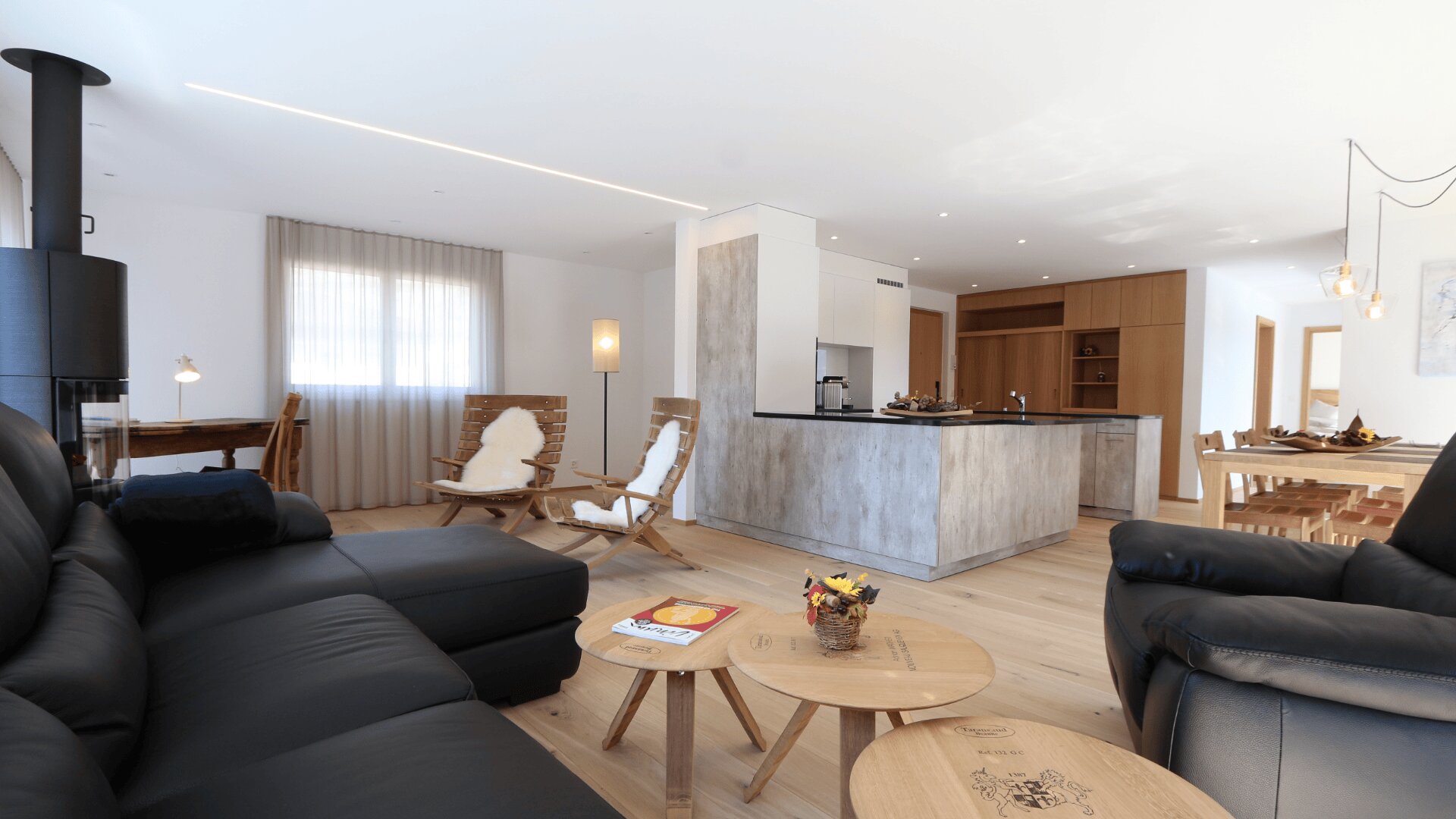 Ein moderner, offener Wohnbereich mit schwarzen Ledersofas, runden Holztischen, einer Kücheninsel, gemütlichen Sesseln mit Fellüberwürfen, einem Esstisch aus Holz und großen Fenstern, die natürliches Licht hereinlassen.