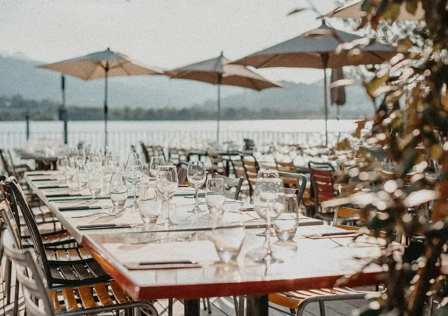 Die Tische im Außenbereich des Restaurants sind mit Gläsern, Tellern und Besteck eingedeckt und stehen in Reihen unter großen Sonnenschirmen, mit Blick auf das Wasser und die Berge im Hintergrund an einem sonnigen Tag.