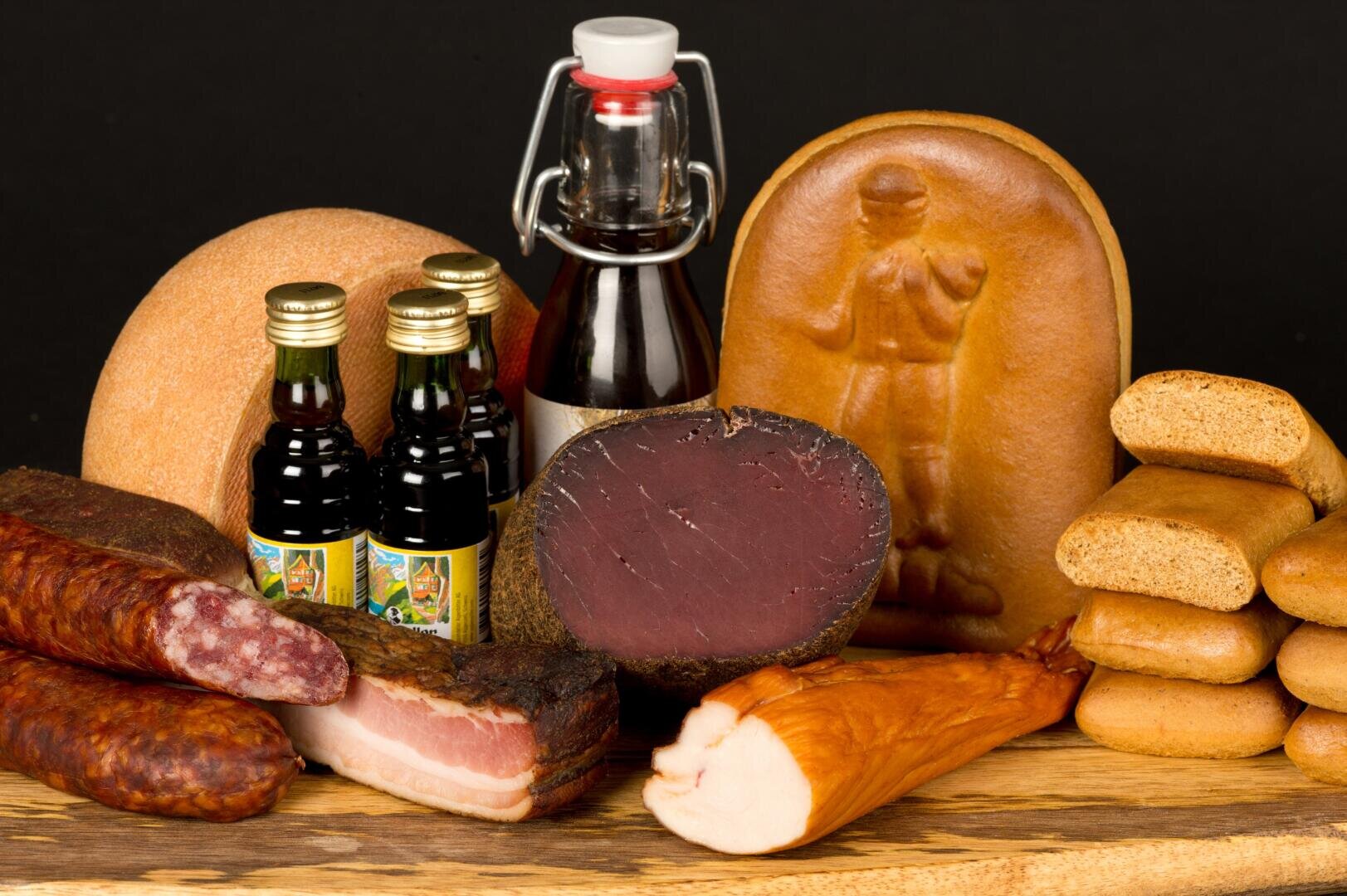 Auf einer Holzfläche vor schwarzem Hintergrund sind verschiedene traditionelle Räucherwaren, Würste, Speck, Flaschen mit dunkler Flüssigkeit und runde, geprägte Brote angeordnet.