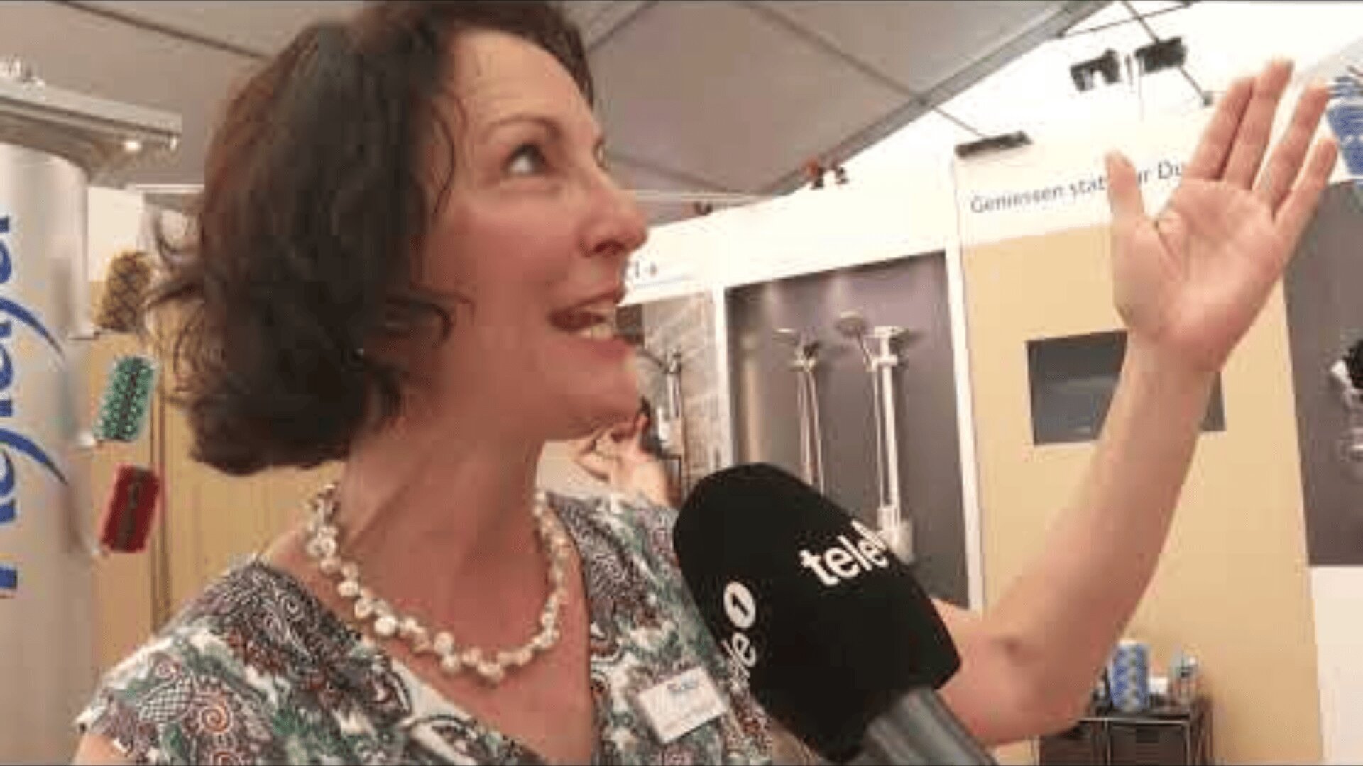 Eine Frau mit kurzen dunklen Haaren und einer Perlenkette spricht enthusiastisch und gestikuliert mit der Hand. Sie wird mit einem Mikrofon mit der Aufschrift tele1 bei einer Veranstaltung oder Ausstellung in einem Gebäude interviewt.