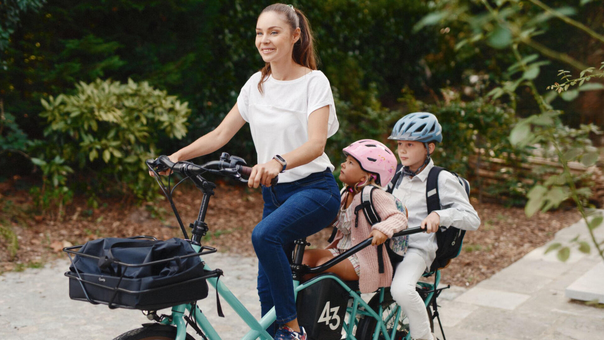 Eine Frau fährt auf einem Fahrrad, hinter ihr sitzen zwei Kinder, die beide Helme und Rucksäcke tragen. Die Gruppe befindet sich im Freien auf einem Weg, der von Grün umgeben ist.