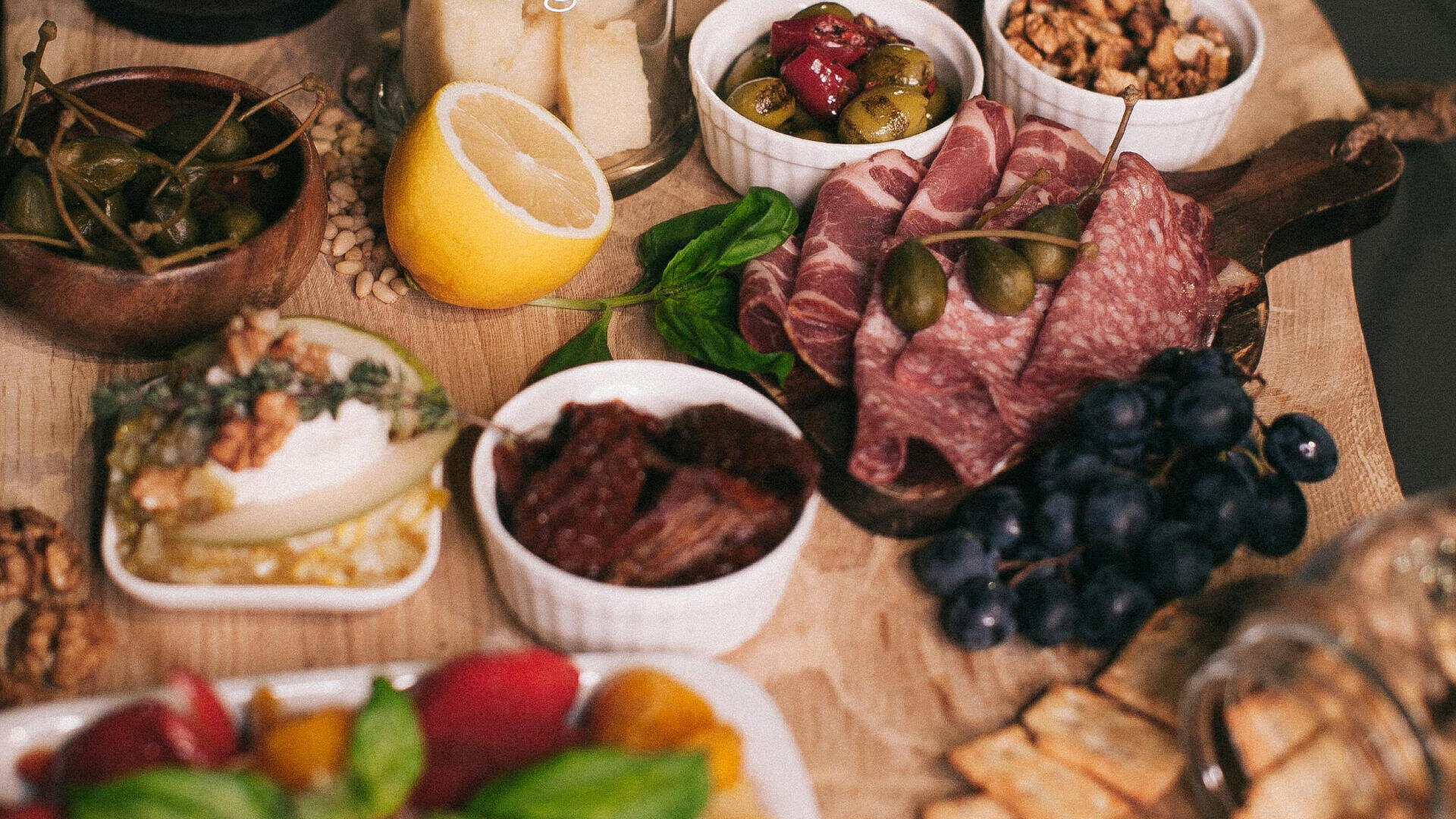 Ein Holzbrett, gefüllt mit verschiedenen Lebensmitteln, darunter Wurstwaren, Käse, Oliven, Trauben, Cracker, Zitrone, Kapern und kleine Schälchen mit Dips und Nüssen, arrangiert für einen Aufstrich aus Wurstwaren.