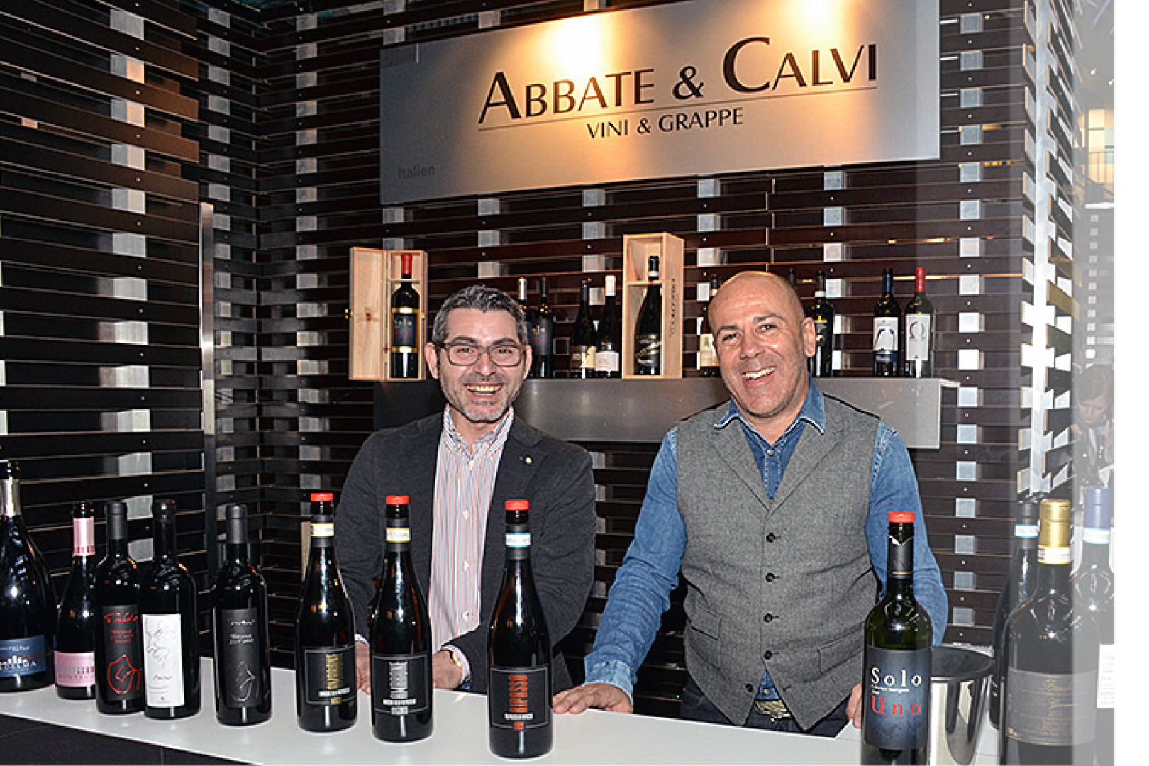Zwei lächelnde Männer stehen hinter einer Theke mit verschiedenen Weinflaschen, über denen ein Schild mit der Aufschrift "Abbate & Calvi Vini e Grappe" hängt. Regale mit Flaschen säumen die dunkel gestreifte Wand hinter ihnen.