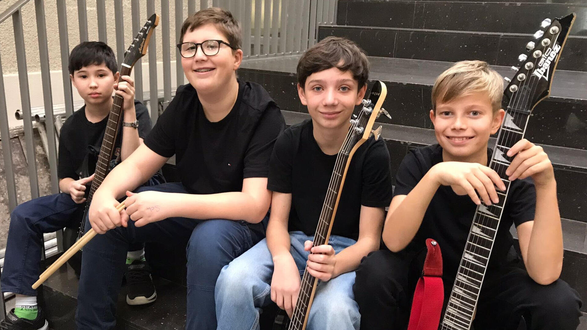 Vier Jungen sitzen auf einer Treppe, jeder hält ein Musikinstrument in der Hand. Zwei halten E-Gitarren, einer hat eine Bassgitarre und ein anderer hält Schlagzeugstöcke. Sie tragen alle schwarze Hemden und lächeln in die Kamera.