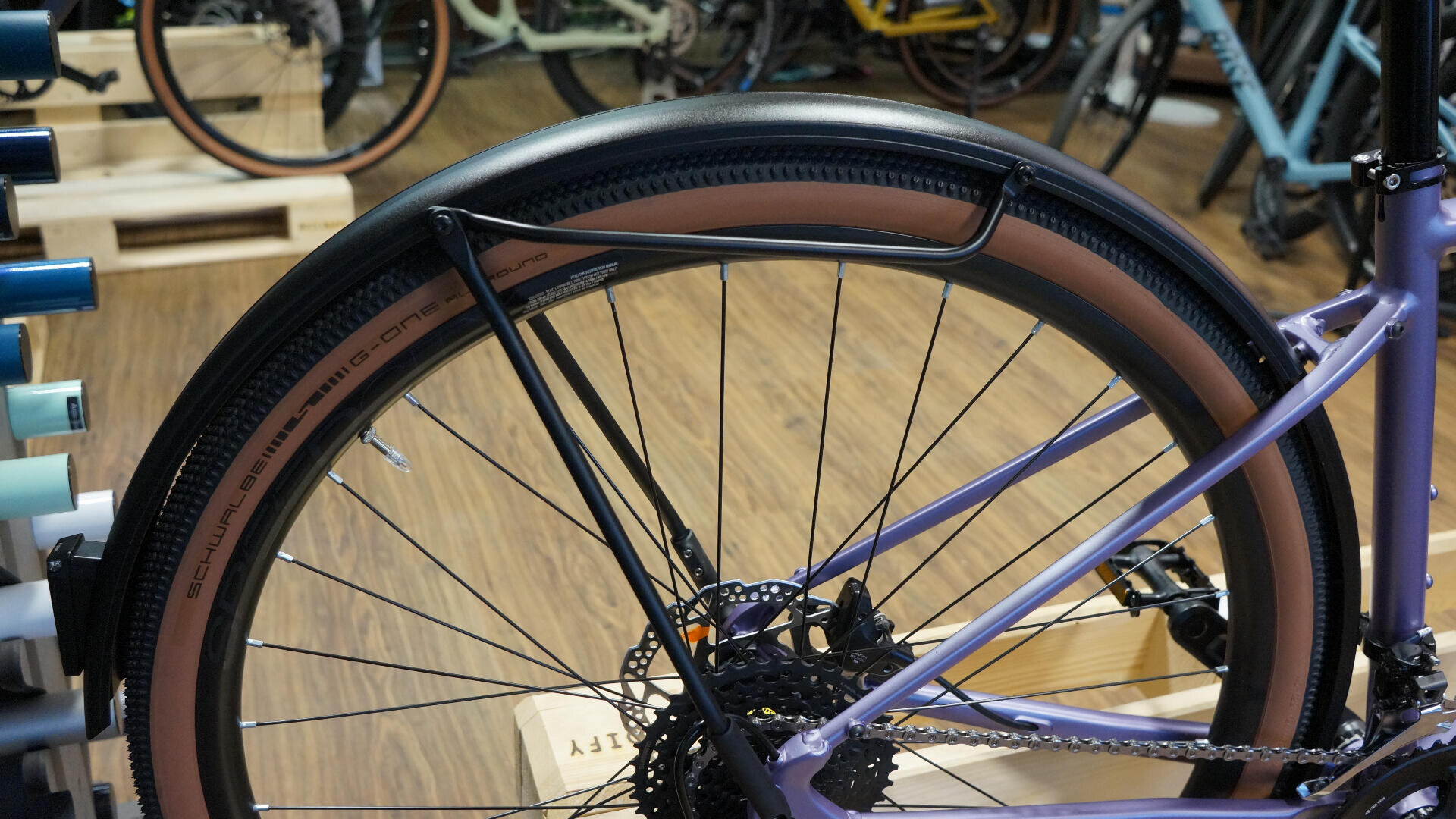Eine Nahaufnahme des Hinterrads eines Fahrrads mit einem hellbraunen Schwalbe-Reifen, einer Scheibenbremsscheibe und einem schwarzen Metallschutzblech vor einem Holzboden und anderen Fahrrädern im Hintergrund.