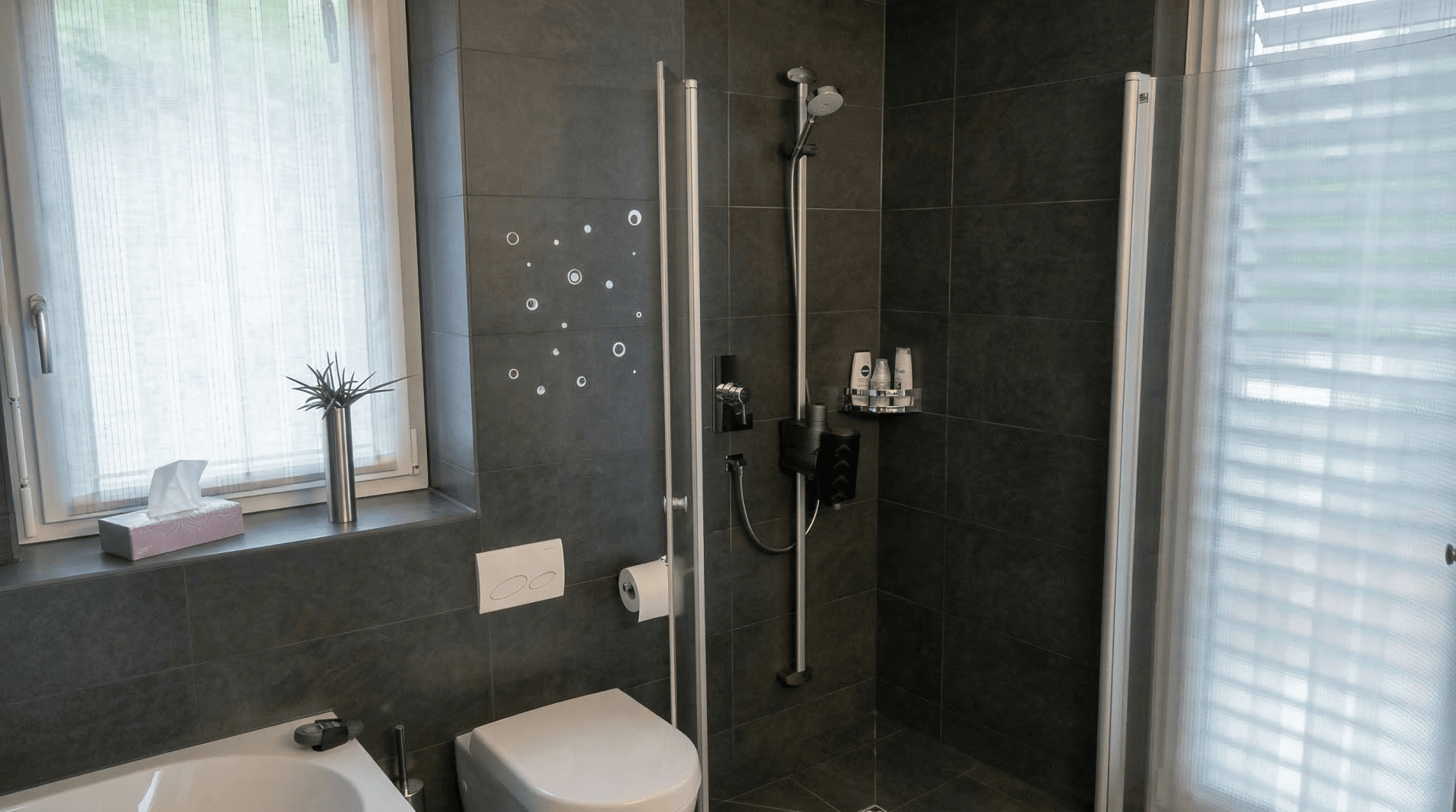 Modernes Badezimmer mit dunkelgrauen Fliesen, einer wandmontierten Toilette, einer Dusche mit Glastüren und einem kleinen Regal mit Toilettenartikeln. Neben der Toilette sind ein Fenster mit durchsichtigen Vorhängen und eine kleine Pflanze in einer Vase zu sehen.