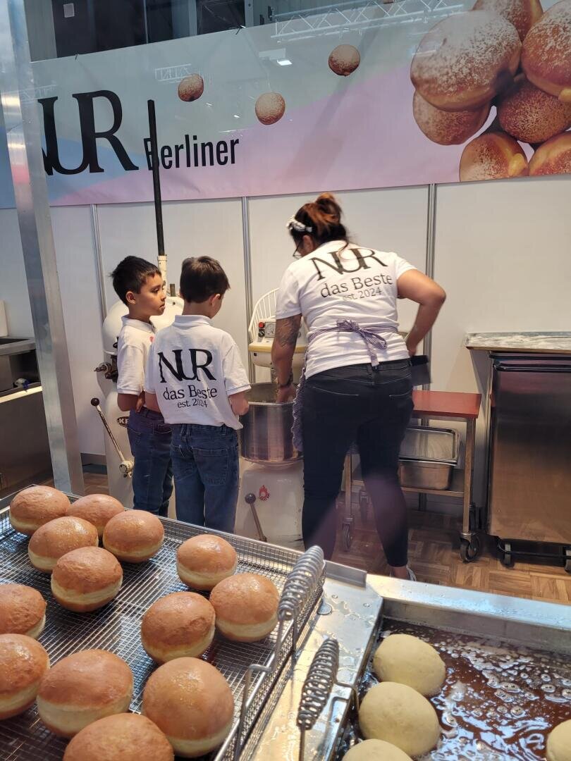 Drei Kinder und eine Frau, die alle NR das Beste-Shirts tragen, stehen an einer Fritteuse an einem Stand mit Tabletts mit Berliner Krapfen. Die Szene scheint auf einer Veranstaltung oder einem Markt zu spielen, bei dem es um die Herstellung oder den Verkauf von Gebäck geht.