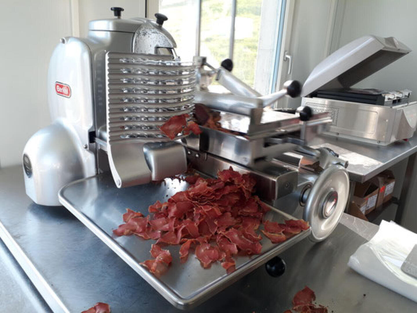 Eine industrielle Fleischschneidemaschine schneidet dünne Scheiben von gepökeltem Fleisch, die sich auf einem Metalltablett darunter stapeln. Die Maschine steht auf einem Tisch aus rostfreiem Stahl in einem gut beleuchteten Raum mit großen Fenstern.