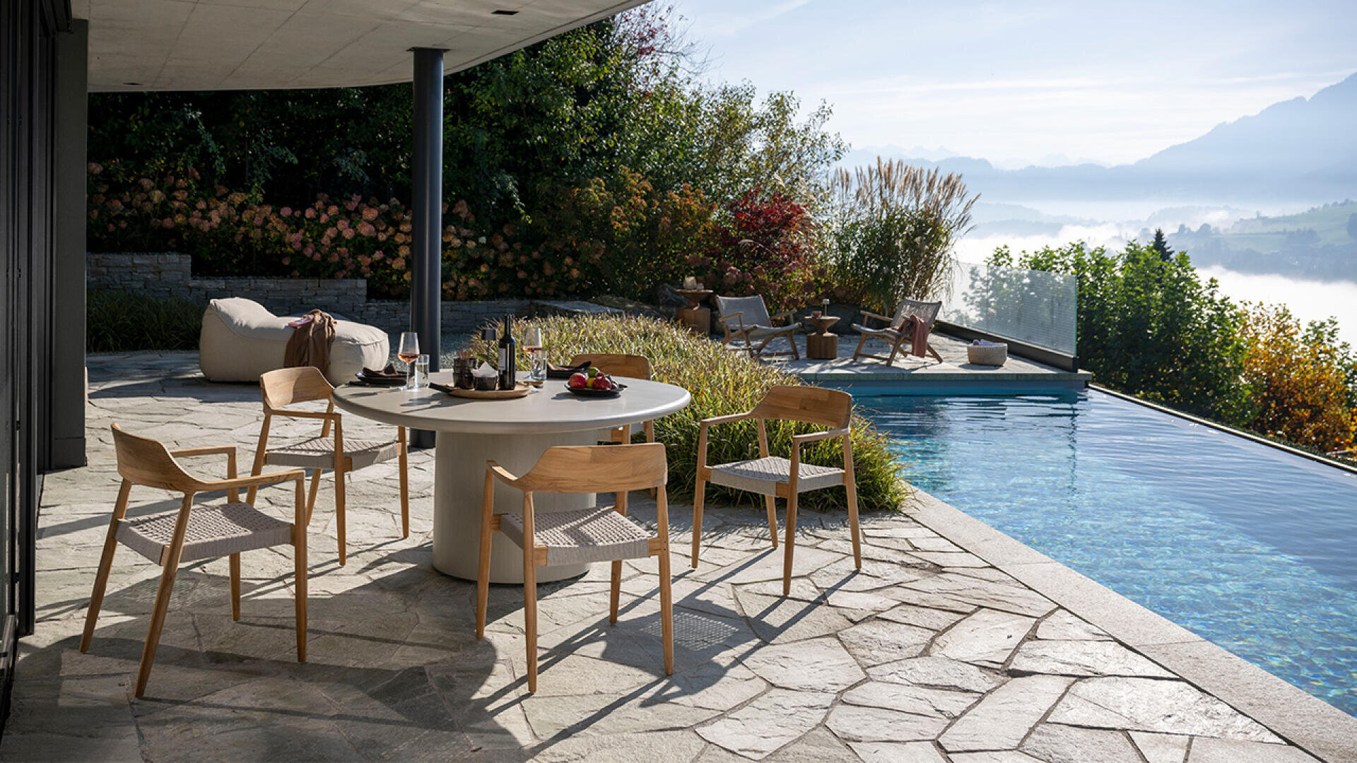 Eine moderne Terrasse mit einem runden Esstisch und vier Holzstühlen blickt auf einen Infinity-Pool und üppiges Grün. Die Leute entspannen sich am Pool, während im Hintergrund neblige Hügel und Berge zu sehen sind.