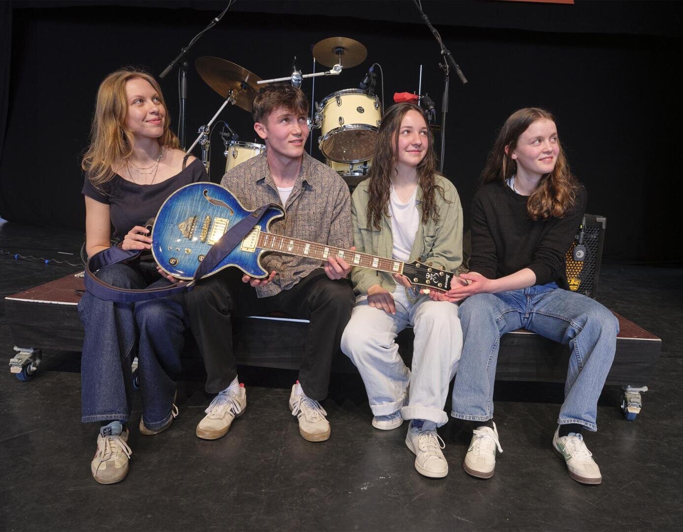 Vier junge Erwachsene sitzen zusammen auf einer Bühne vor einem Schlagzeug. Eine Person hält eine blaue E-Gitarre. Alle sind leger gekleidet, lächeln und blicken leicht aus der Kamera.