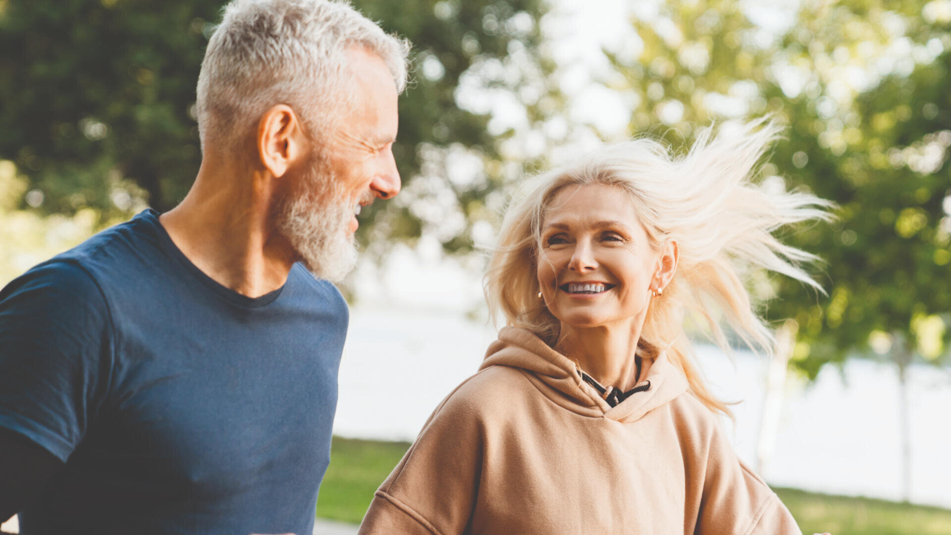 Ein lächelnder älterer Mann und eine Frau joggen an einem sonnigen Tag im Freien und schauen sich an. Im Hintergrund sind grüne Bäume und eine helle, natürliche Umgebung zu sehen.