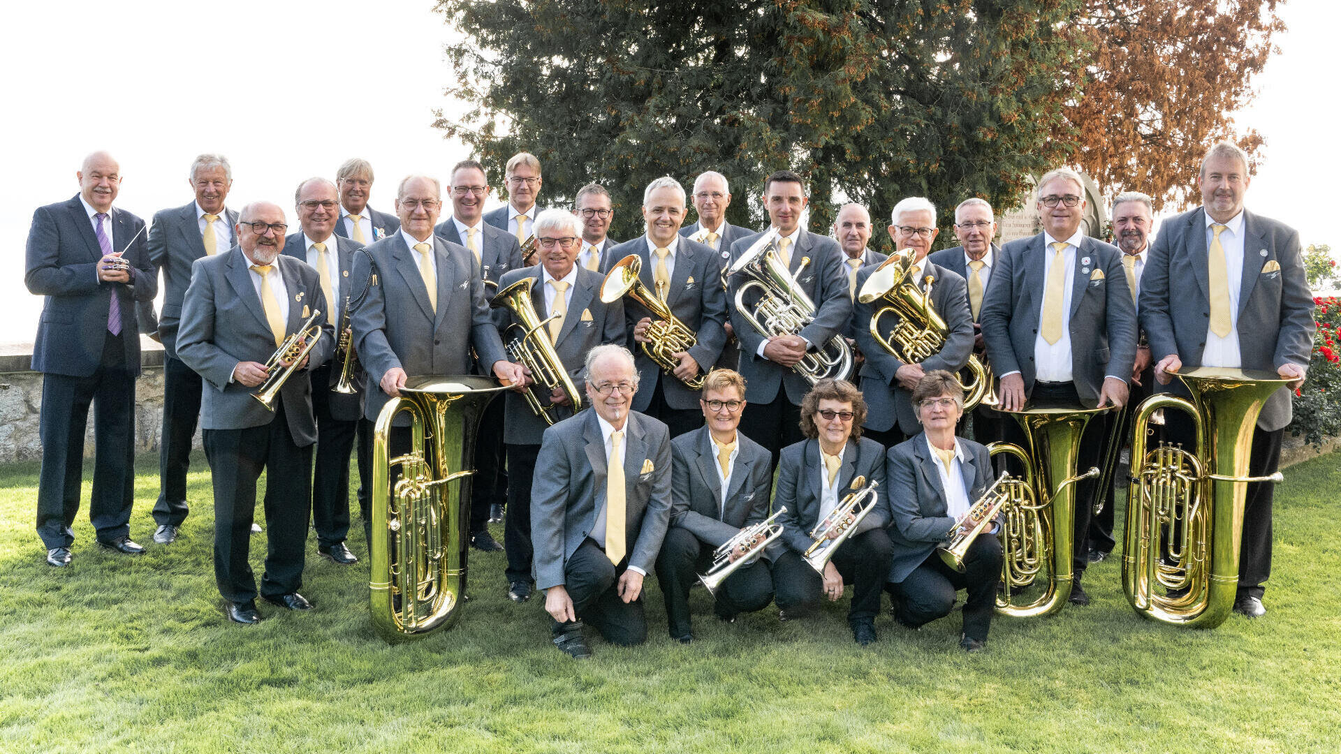Eine Gruppe von Musikern in grauen Anzügen mit gelben Krawatten posiert im Freien auf einer Wiese und hält Blechblasinstrumente wie Tuben, Trompeten und Posaunen. Im Hintergrund sind Bäume und eine Steinmauer zu sehen.