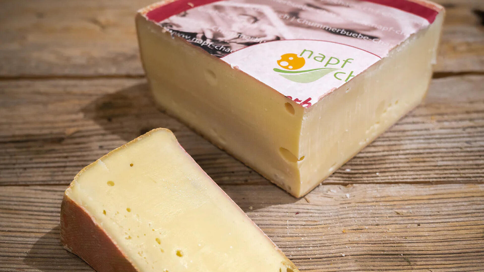 Ein Stück Napf-Chäs-Käse liegt auf einer Holzunterlage. Im Vordergrund ist eine kleinere Scheibe zu sehen.