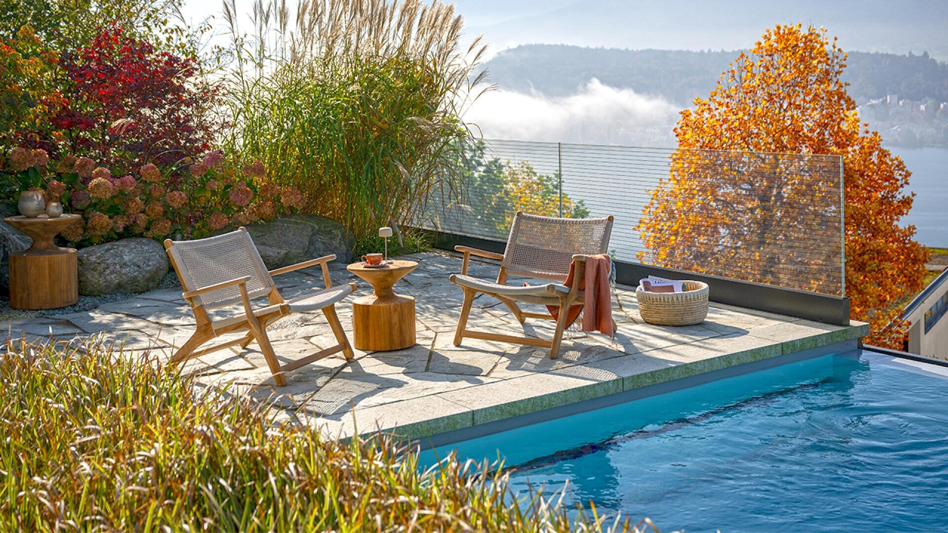 Zwei Holzstühle und ein kleiner Tisch stehen auf einer Steinterrasse neben dem Pool, umgeben von bunten Herbstpflanzen und Bäumen, mit Blick auf ein Gewässer und die nebligen Berge im Hintergrund.