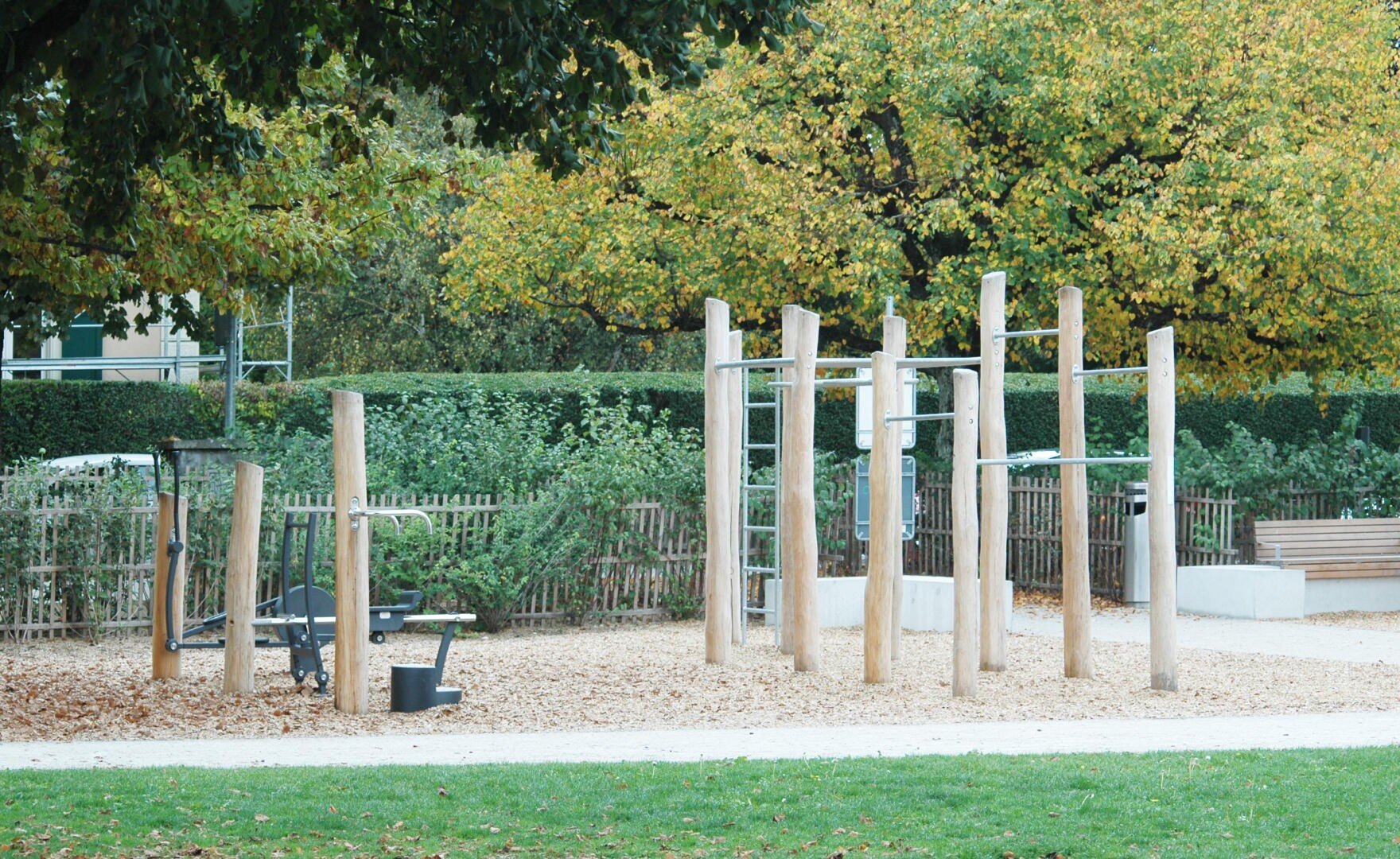 Ein Spielplatz im Freien mit hölzernen Kletterstangen, Reckstangen und Fitnessgeräten, alles auf einer Holzspanfläche mit grünem Gras und Bäumen mit Herbstlaub im Hintergrund.