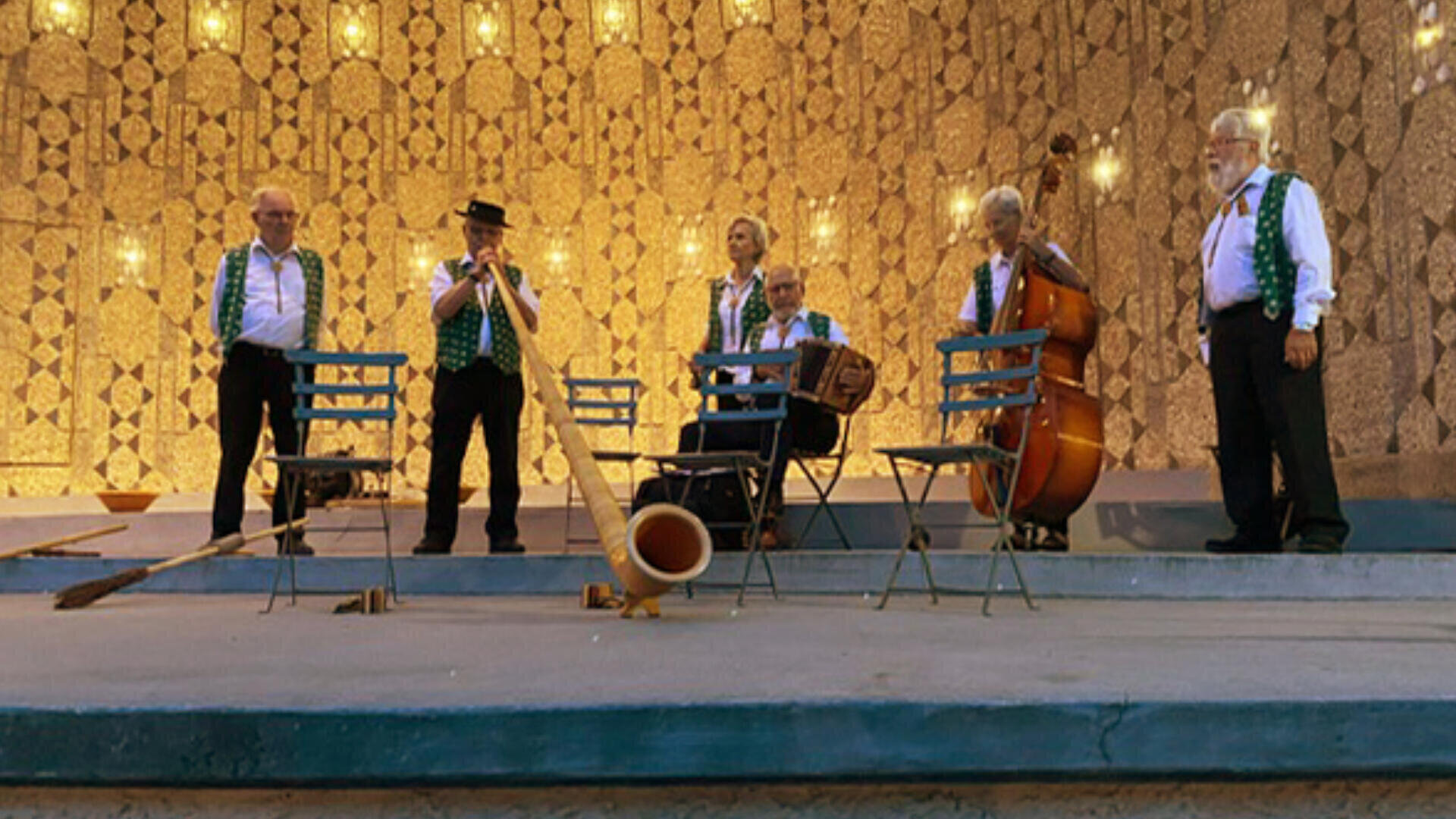 Eine Gruppe von sechs Musikern in traditioneller Kleidung spielt auf der Bühne mit Instrumenten wie Alphorn, Akkordeon und Kontrabass vor einer Kulisse aus Hängelampen und gemusterten Wänden.