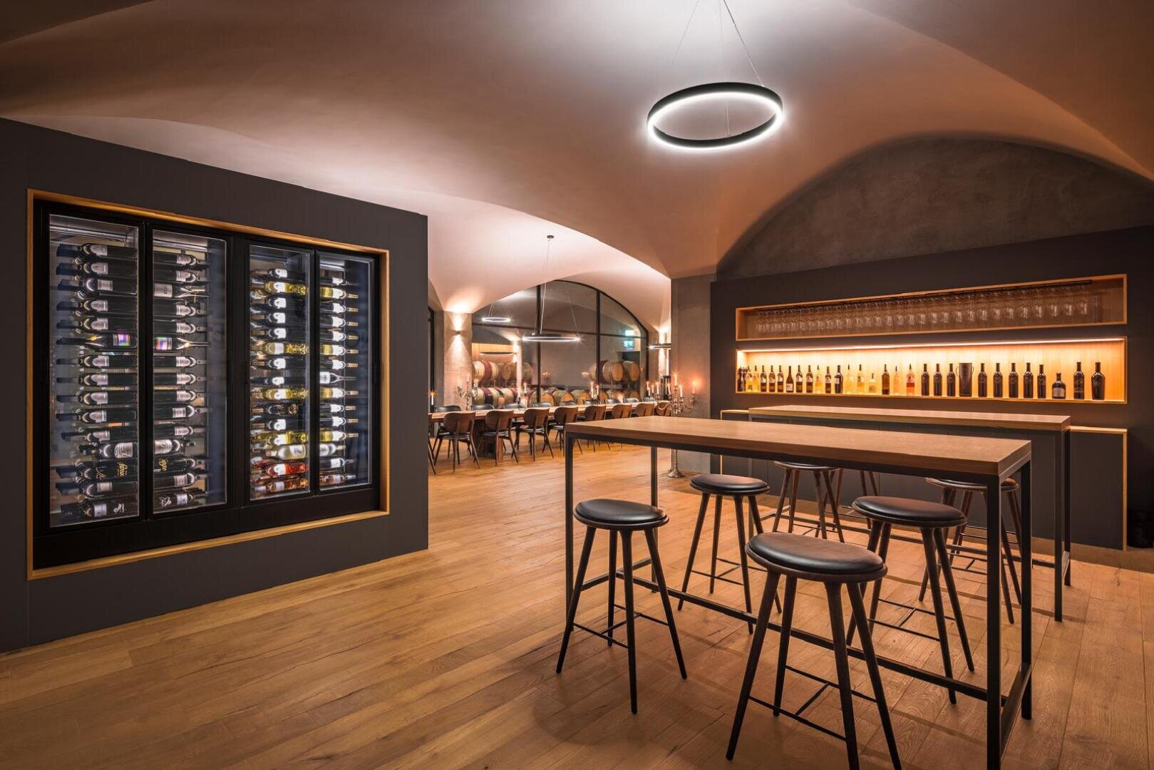 Eine moderne Weinbar mit Holzböden, einem hohen Tisch und Hockern, einer Wand mit Weinflaschen, einem eleganten Weinkühlschrank, Hängeleuchten und Regalen mit Gläsern hinter der Bar. Der Raum hat gewölbte Decken und ein warmes Ambiente.