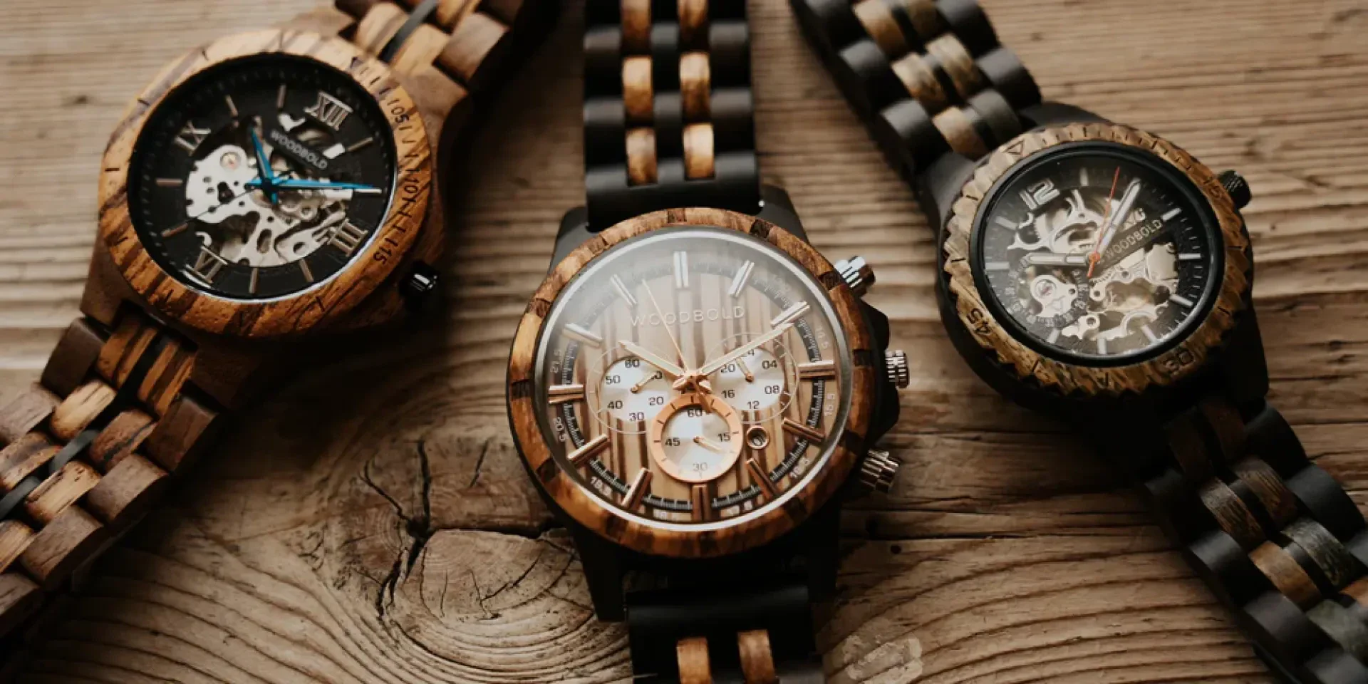 Drei hölzerne Armbanduhren mit freiliegenden mechanischen Zahnrädern sind auf einer rustikalen Holzoberfläche ausgestellt. Die Uhren weisen eine Mischung aus hellen und dunklen Holzbändern und komplizierten, offenen Zifferblättern auf, die ihr Innenleben zeigen.