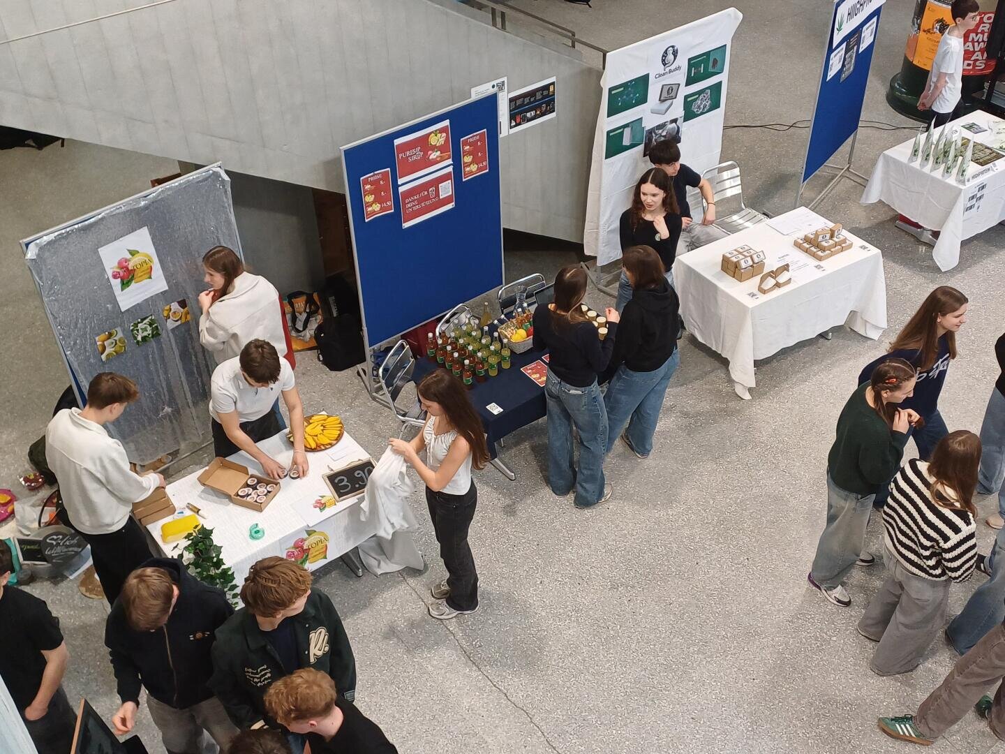 Eine Gruppe von Menschen sitzt an mehreren Tischen mit Speisen, Getränken und Auslagen, die in einem Veranstaltungsraum, möglicherweise einer Messe oder Ausstellung, aufgestellt sind. Einige verkaufen oder präsentieren Gegenstände, während andere sich umsehen oder unterhalten.
