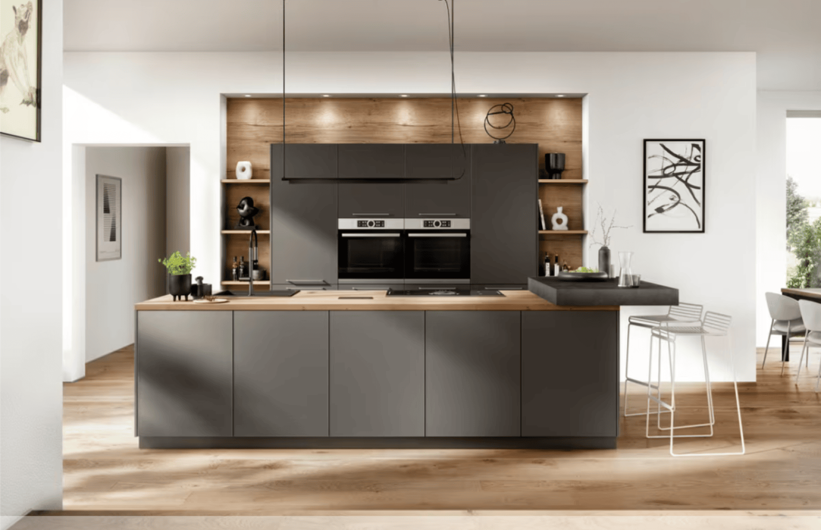 Moderne Küche mit mattgrauen Schränken, Einbaubackofen, Arbeitsplatte aus Holz, offenen Regalen mit Dekorationsartikeln, Barhockern an einem erhöhten Tresen und hellem Holzfußboden in einem hellen, offenen Raum.