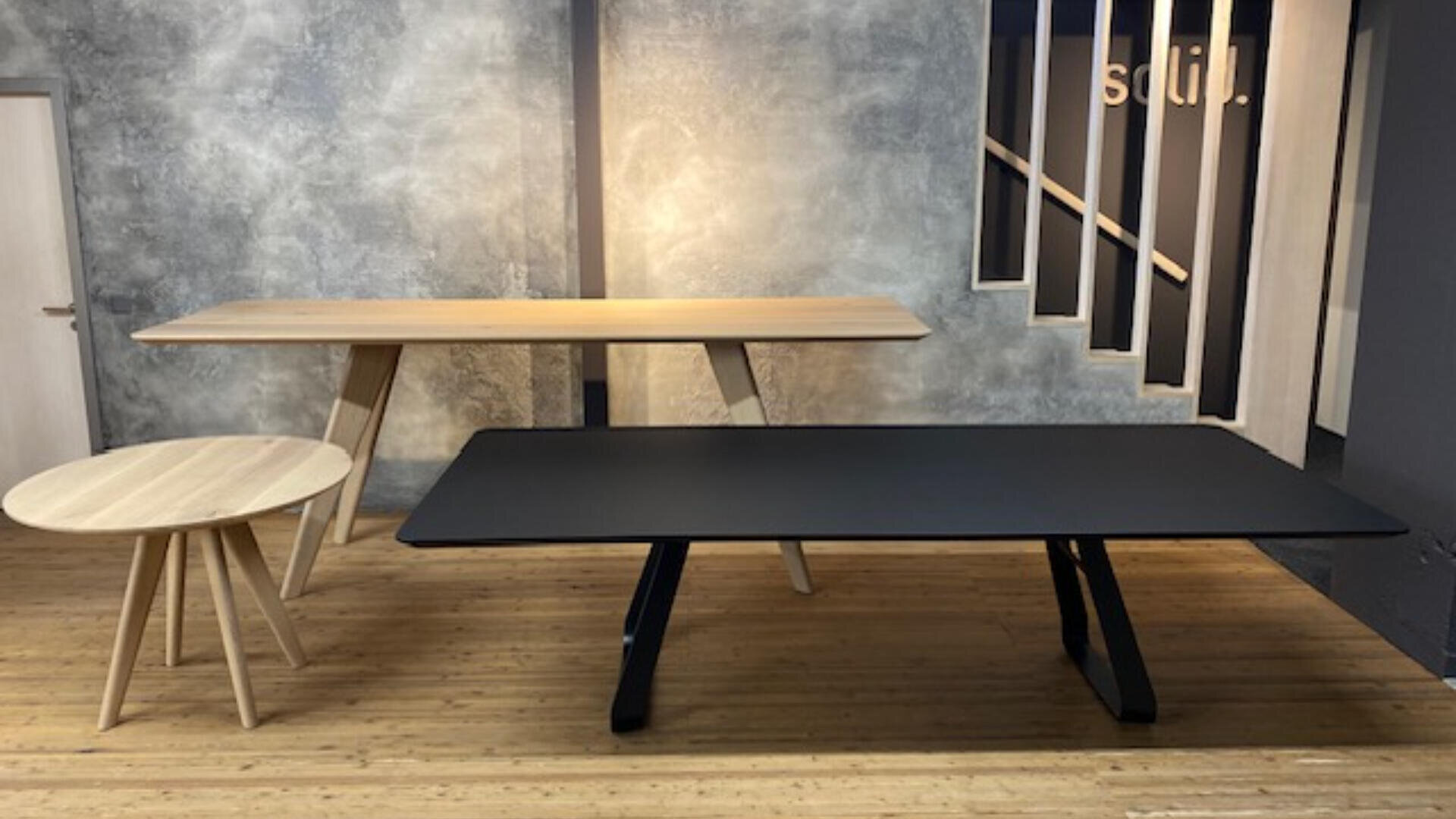 Drei moderne Tische - ein runder heller Holztisch, ein rechteckiger heller Holztisch und ein rechteckiger schwarzer Tisch - stehen auf einem Holzboden vor einer grau strukturierten Wand mit einer Treppe und einer Tür im Hintergrund.