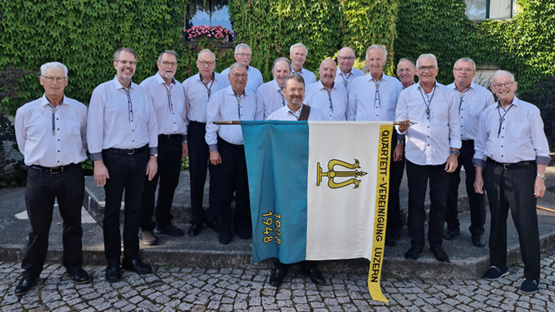 Eine Gruppe von Männern in passenden weissen Hemden und dunklen Hosen steht draussen auf Steinstufen und hält eine Fahne mit der Aufschrift Quartett-Vereinigung Luzern 1948.