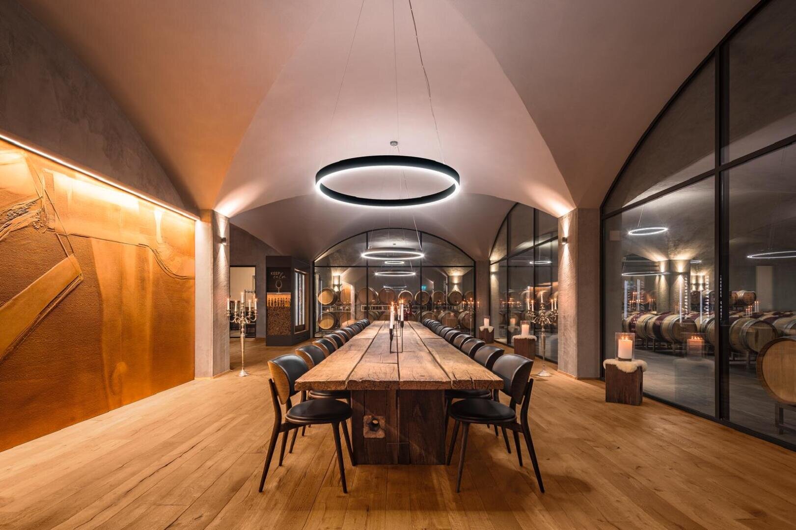 Ein moderner, eleganter Verkostungsraum mit einem langen Holztisch, schwarzen Stühlen, einer gewölbten Decke, runden Beleuchtungskörpern und Glaswänden, die den Blick auf die Weinfässer im angrenzenden Keller freigeben. Die warme Beleuchtung schafft ein gemütliches Ambiente.