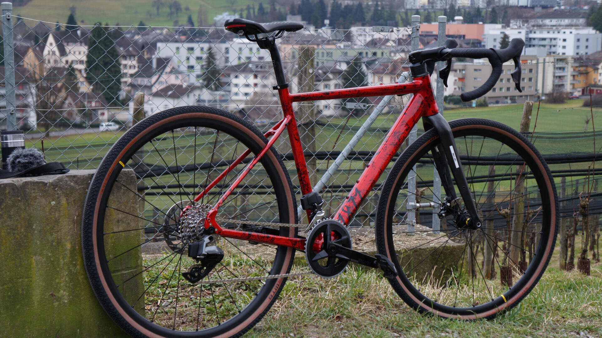 Ein rotes und graues Schotterfahrrad ist auf einer Wiese geparkt, mit einem Zaun und einer Stadtlandschaft aus Gebäuden und Hügeln im Hintergrund.