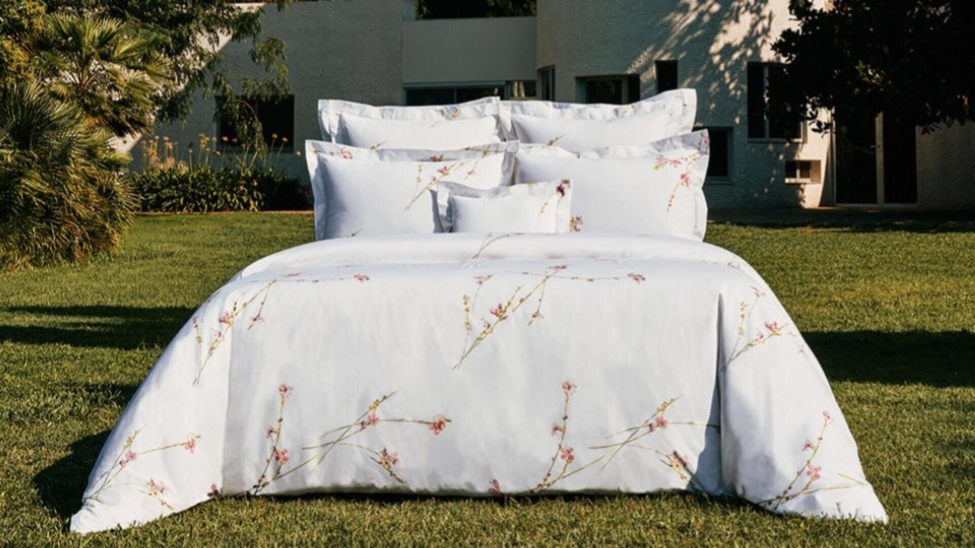 Ein ordentlich gemachtes Bett mit weißem Bettzeug, das mit zarten rosa Blumenmustern verziert ist, steht im Freien auf einer grünen Wiese, mit einem modernen weißen Gebäude und Bäumen im Hintergrund.