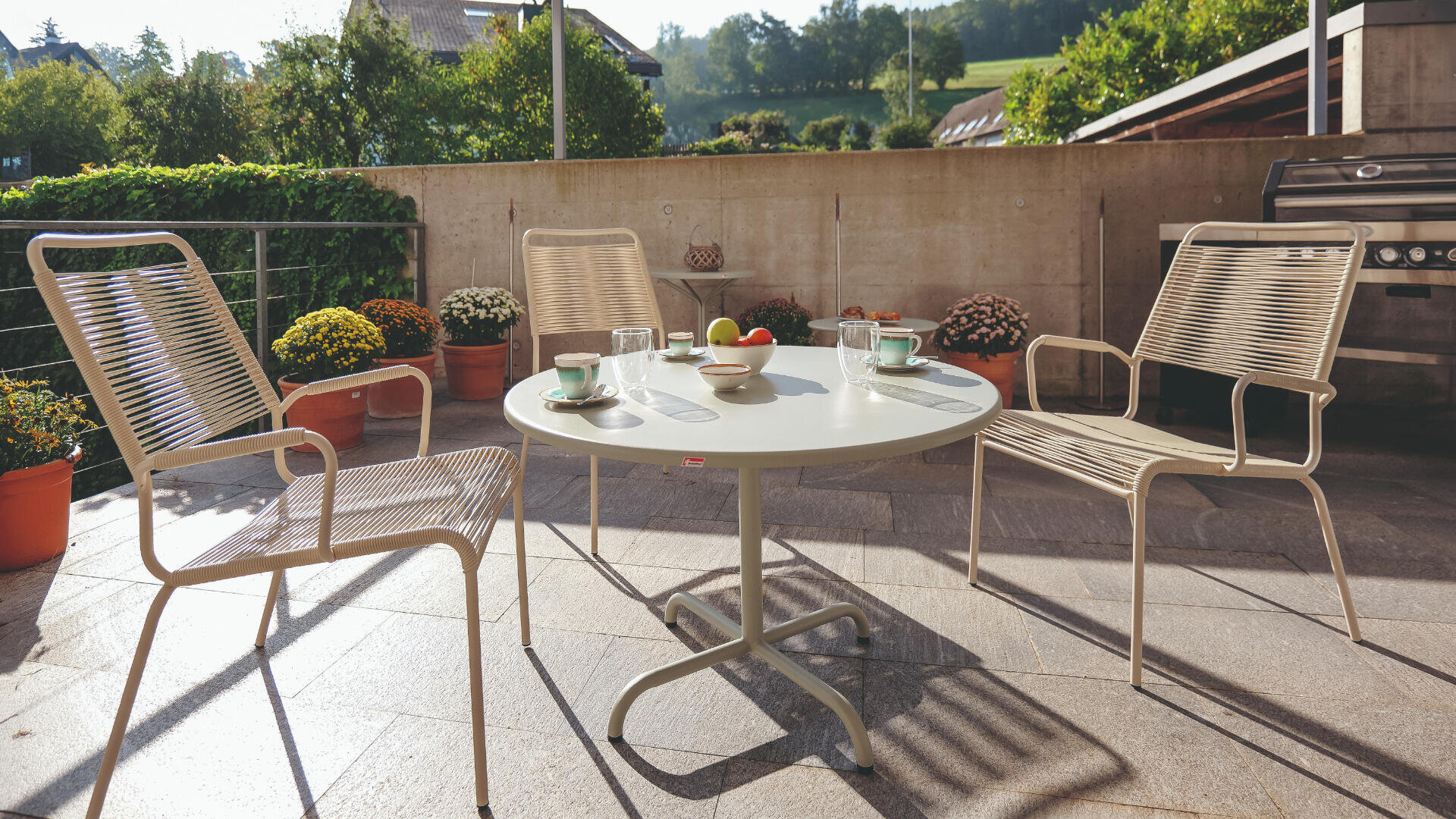 Terrasse im Freien mit drei beigen Stühlen um einen runden weißen Tisch mit Tassen, Gläsern, Obst und einer Wasserflasche. Topfpflanzen säumen den Steinboden; im Hintergrund sind Grünflächen und Hügel zu sehen.