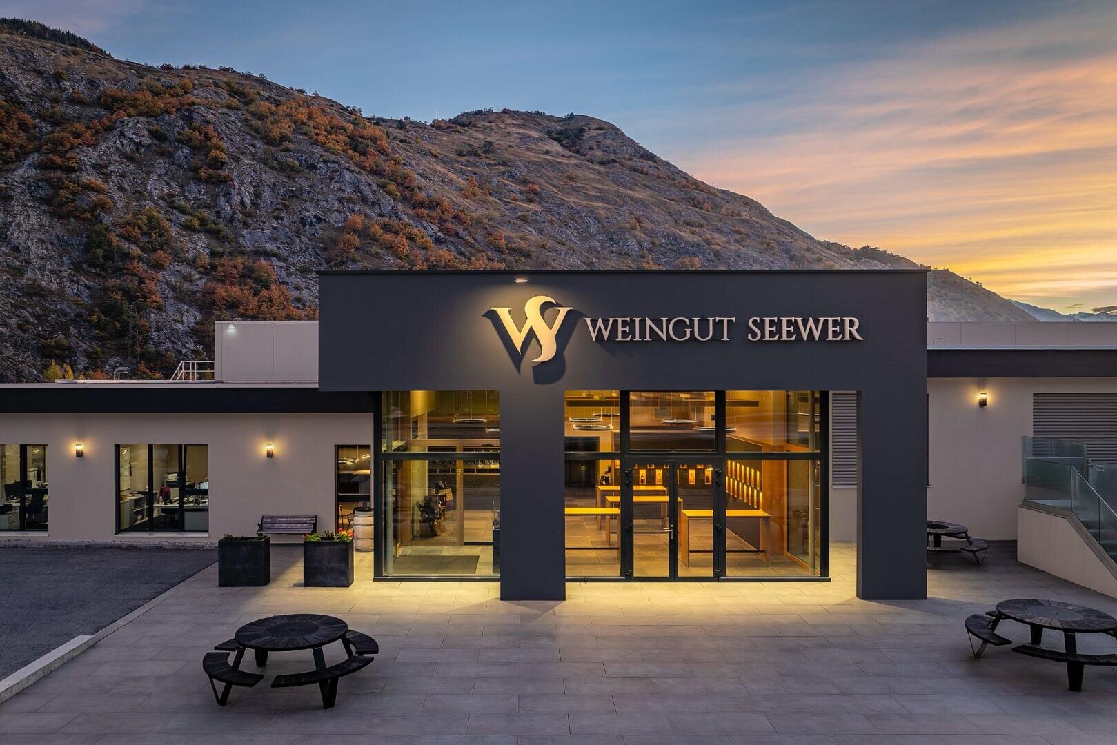 Modernes Kellereigebäude mit der Bezeichnung Weingut Seewer mit großen Glasfenstern, Sitzplätzen im Freien und einer Berglandschaft im Hintergrund bei Sonnenuntergang.