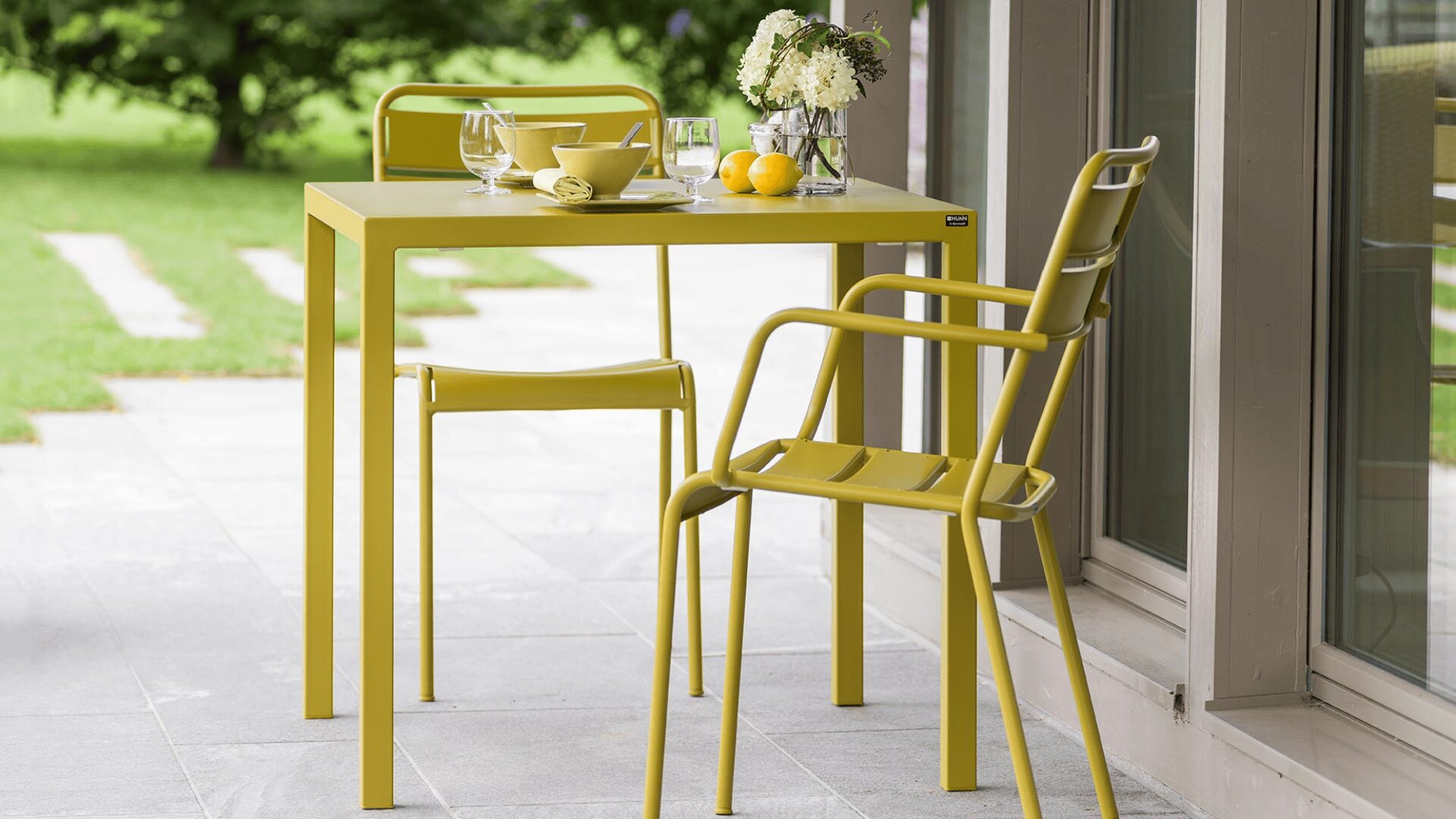 Ein modernes gelbes Outdoor-Essgeschirr mit zwei Stühlen und einem quadratischen Tisch steht auf einer Veranda. Auf dem Tisch stehen Gläser, eine Vase mit Blumen, Zitronen und eine Schale. Im Hintergrund sind grünes Gras und Bäume zu sehen.