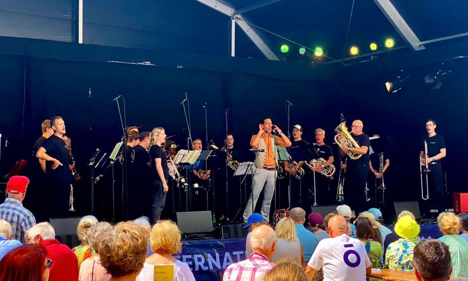 Eine große Blaskapelle spielt auf der Bühne unter einem Zelt, wobei die Musiker verschiedene Blasinstrumente spielen. Ein Publikum in farbenfroher Kleidung beobachtet die Aufführung von unten. Das Bühnenlicht beleuchtet die Musiker.