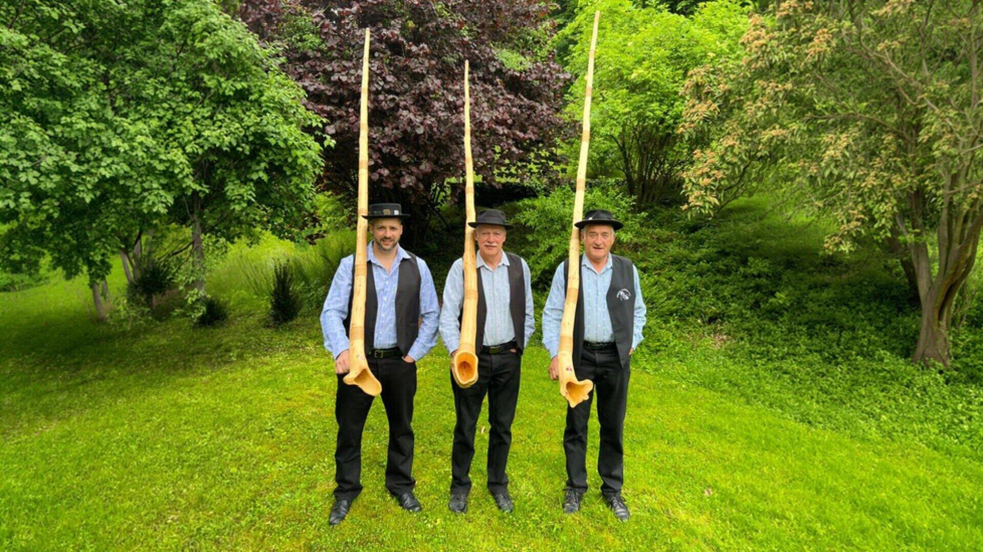 Drei Männer in traditioneller Kleidung stehen im Gras und halten jeweils ein großes Alphorn in die Höhe. Im Hintergrund sind sie von Bäumen und Grünpflanzen umgeben.