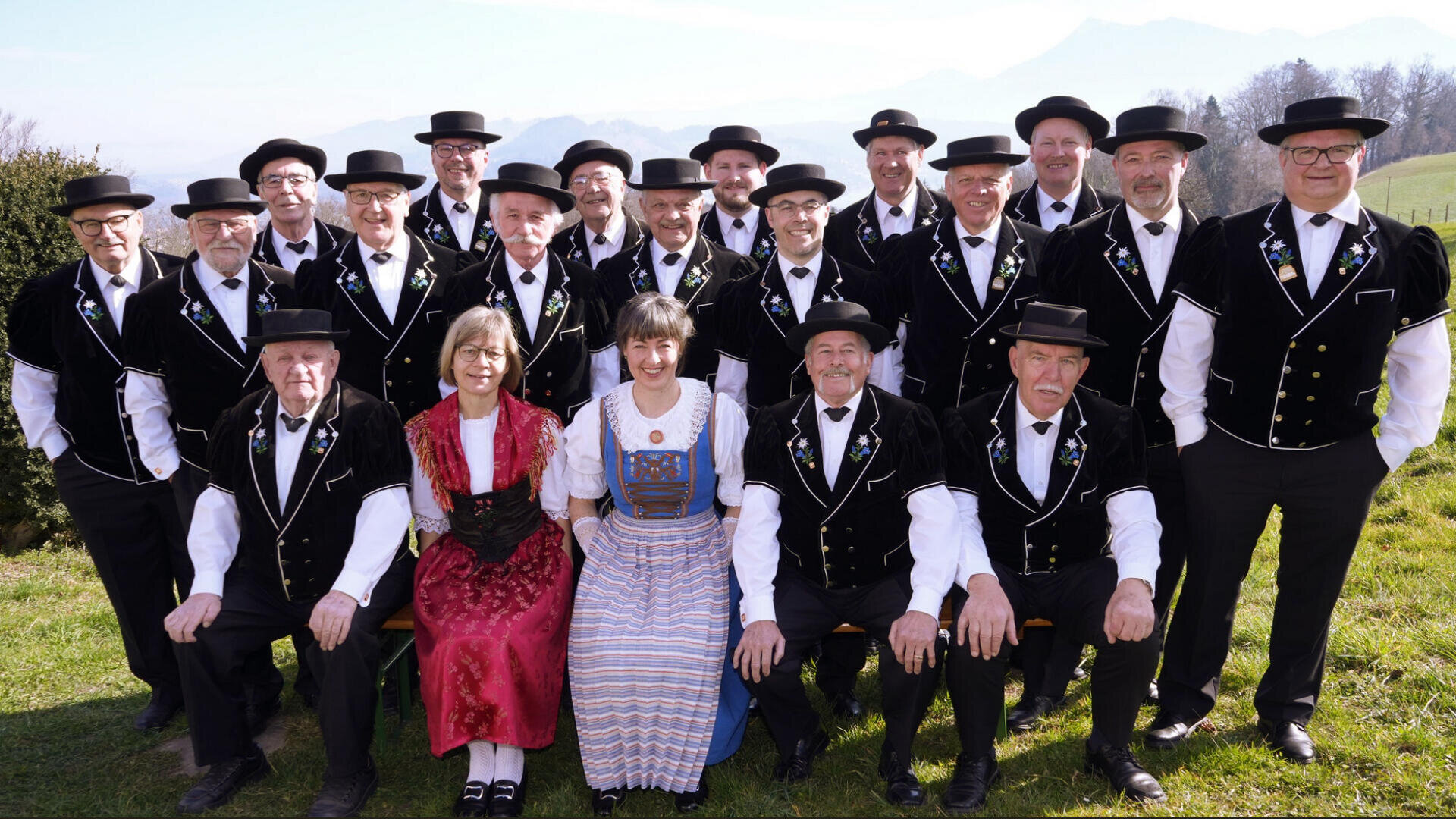 Eine Gruppe von siebzehn Männern in passenden traditionellen schwarzen Anzügen und Hüten posiert im Freien mit zwei Frauen in farbenfrohen traditionellen Kleidern, alle lächeln an einem sonnigen Tag in einer ländlichen Umgebung mit Hügeln im Hintergrund.