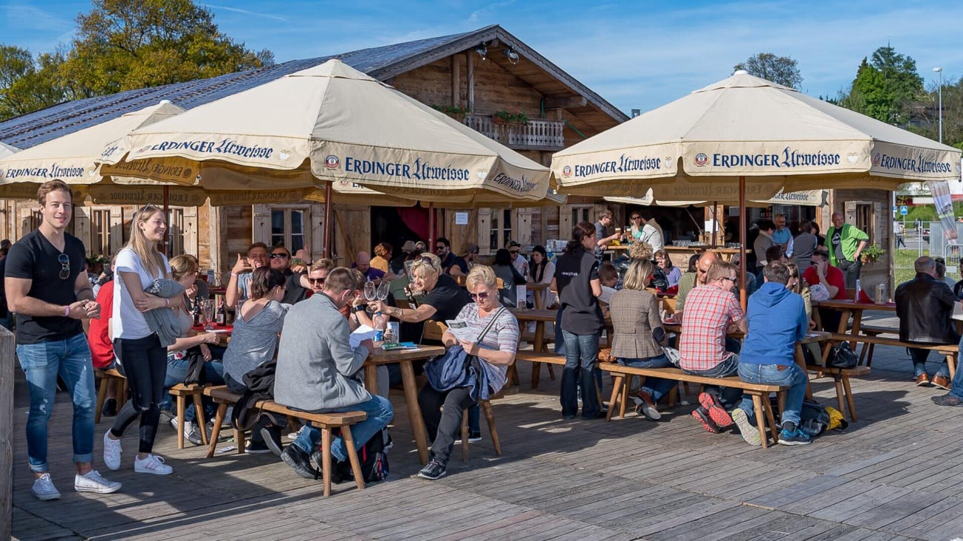 Menschen, die an einem sonnigen Tag an Holztischen im Freien unter großen Erdinger-Schirmen sitzen und sich unterhalten, mit einem rustikalen Holzgebäude und Bäumen im Hintergrund.