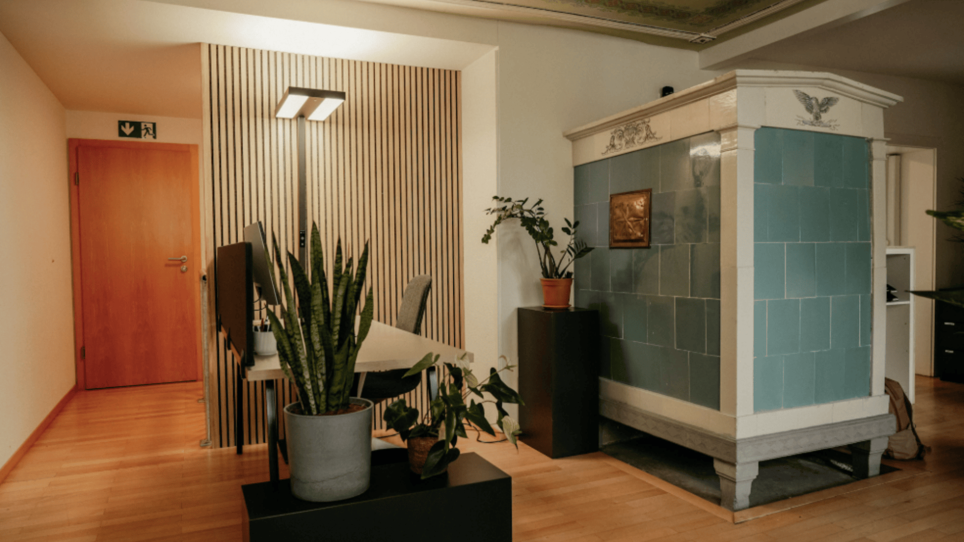 Ein moderner Büroraum mit Holzboden, einem Schreibtisch mit Stühlen, mehreren Topfpflanzen und einem großen Kachelofen mit dekorativen Akzenten an einer Wand. Im Hintergrund befinden sich eine geschlossene Holztür und vertikale Holzlatten.